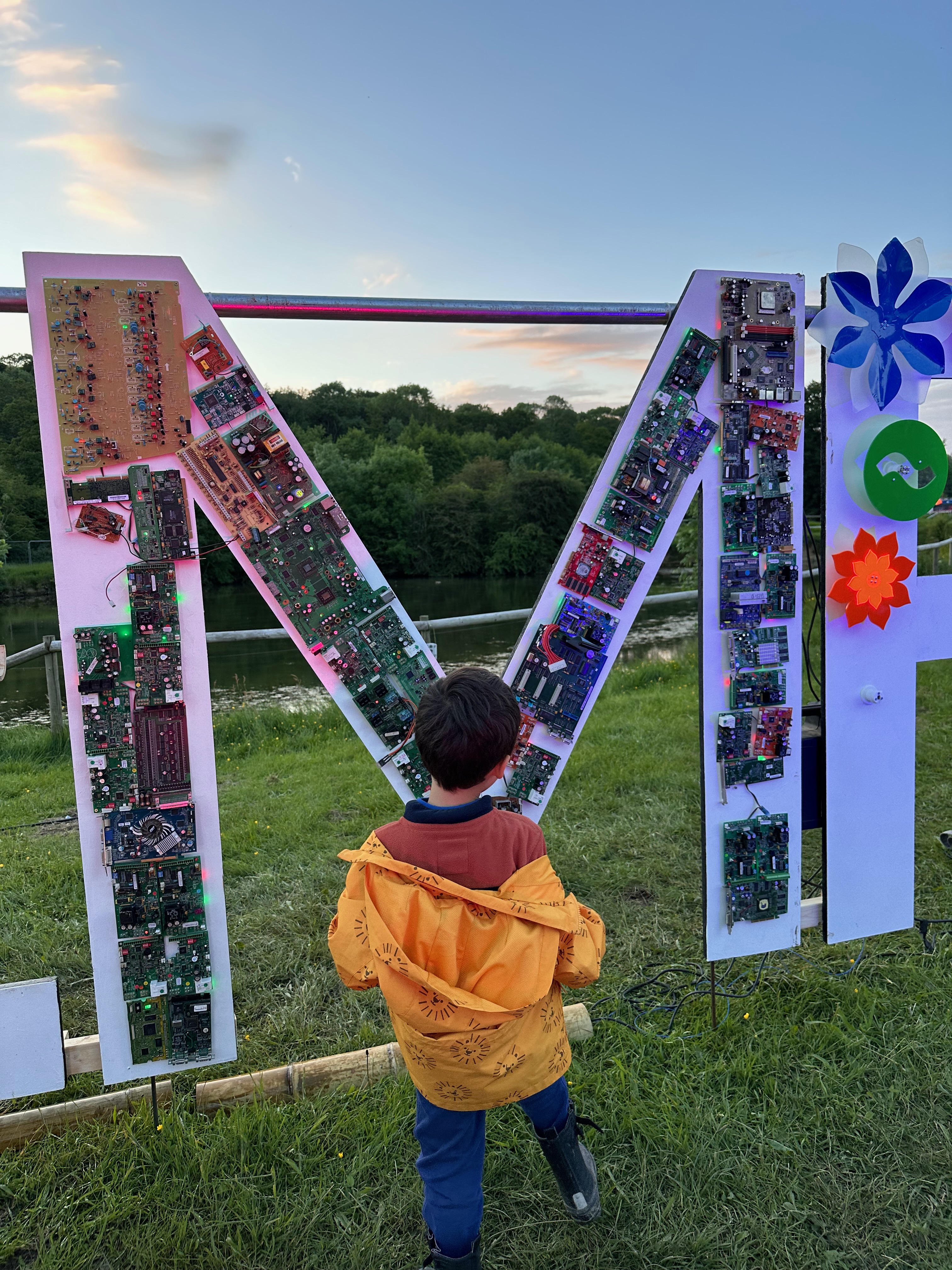 A young boy stands in front of the letter 'M' which is propped up in a field. The 'M' is about twice his height. The letter is white but filled with old circuit boards.