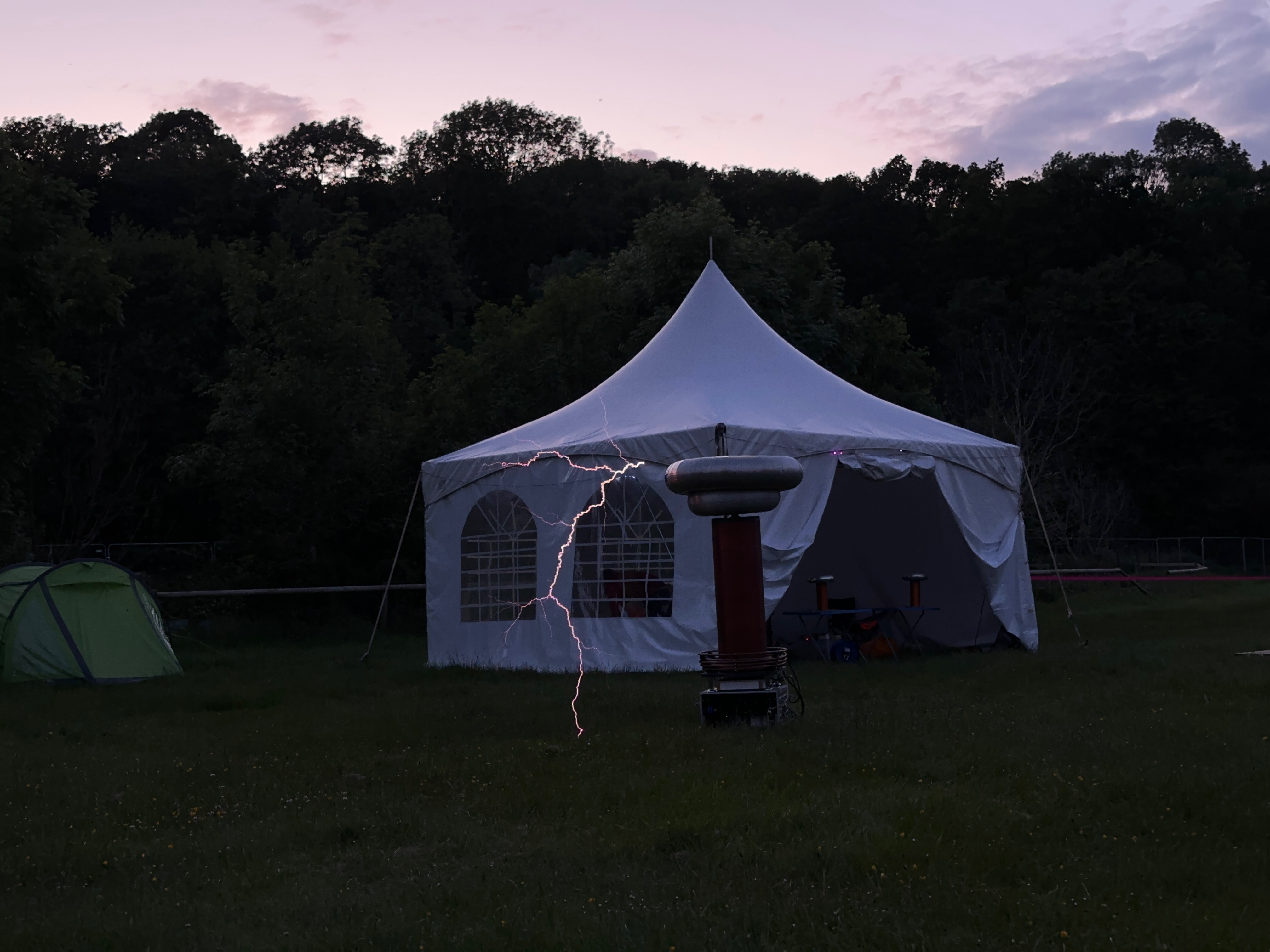 A Tesla coil stands in a field in front of a small marquee tent. A spark leaps from the coil down to the ground.
