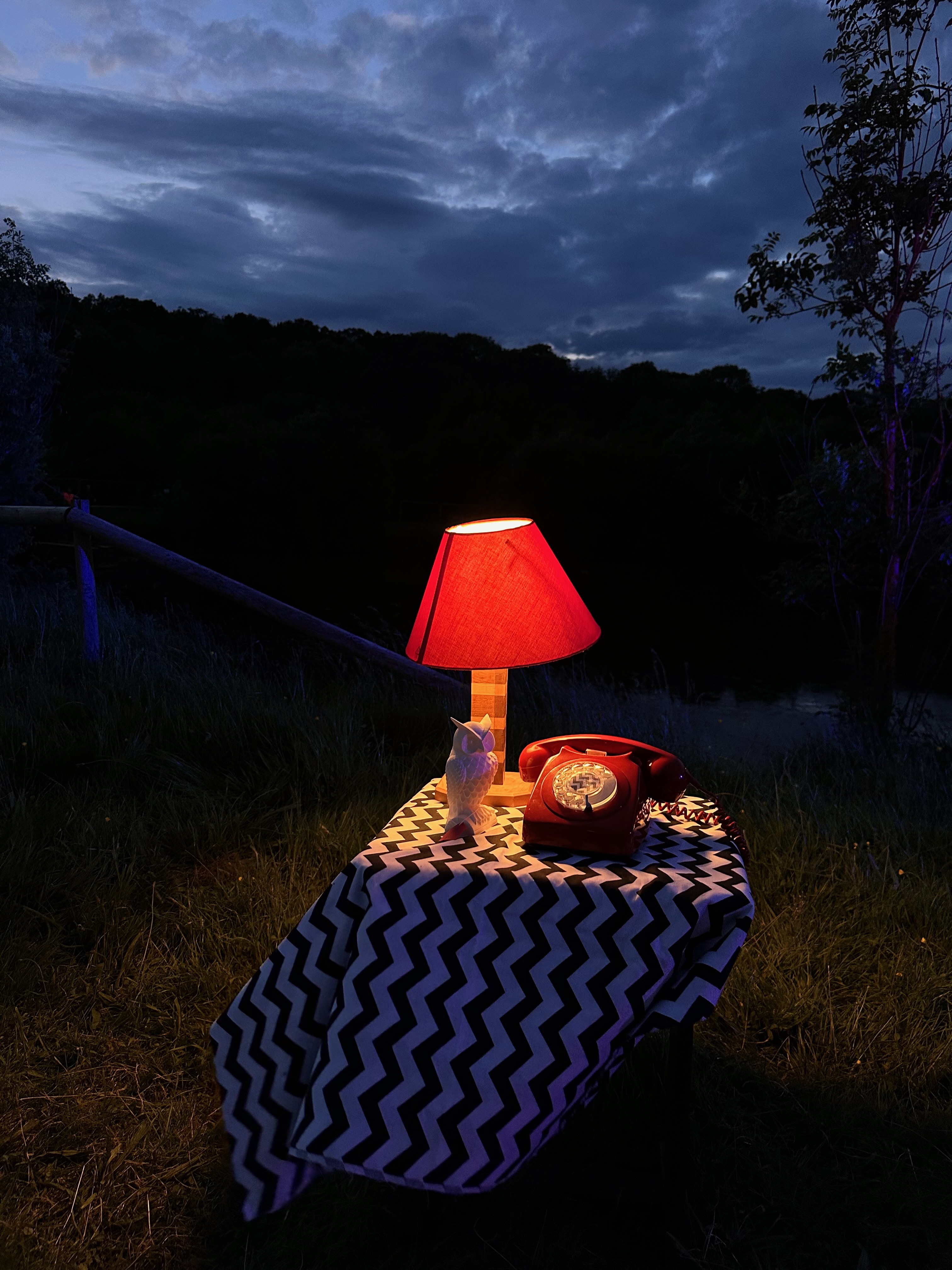 In a dark field, a small square table is covered with a blue and white striped table cloth. On the table stands a red telephone and a white owl. The scene is lit by a small round lamp at the back of the table.
