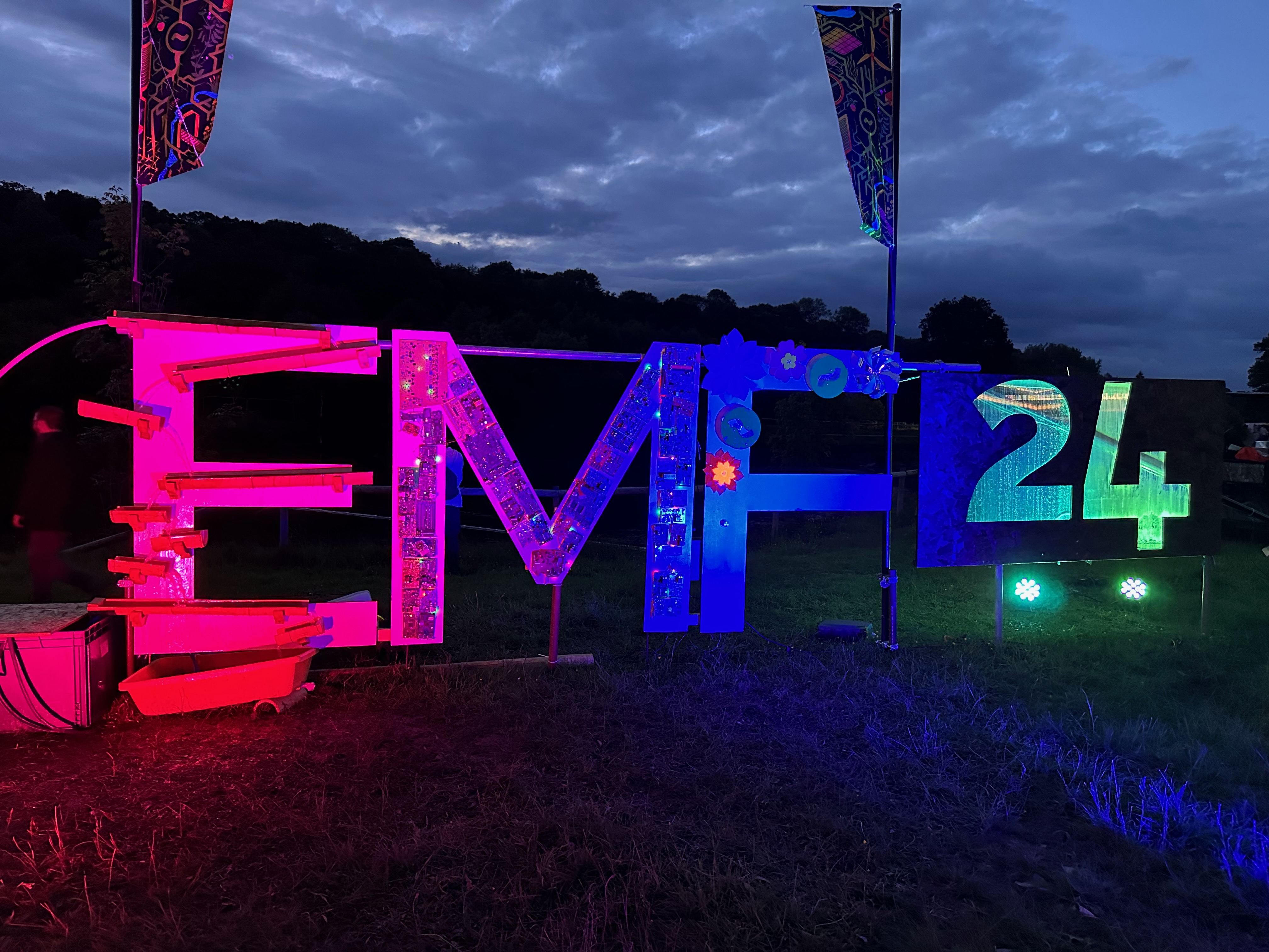 Night time photo of a sign for EMF 24. The letters on the left are lit up pink, fading through blue in the centre and turquise on the right.