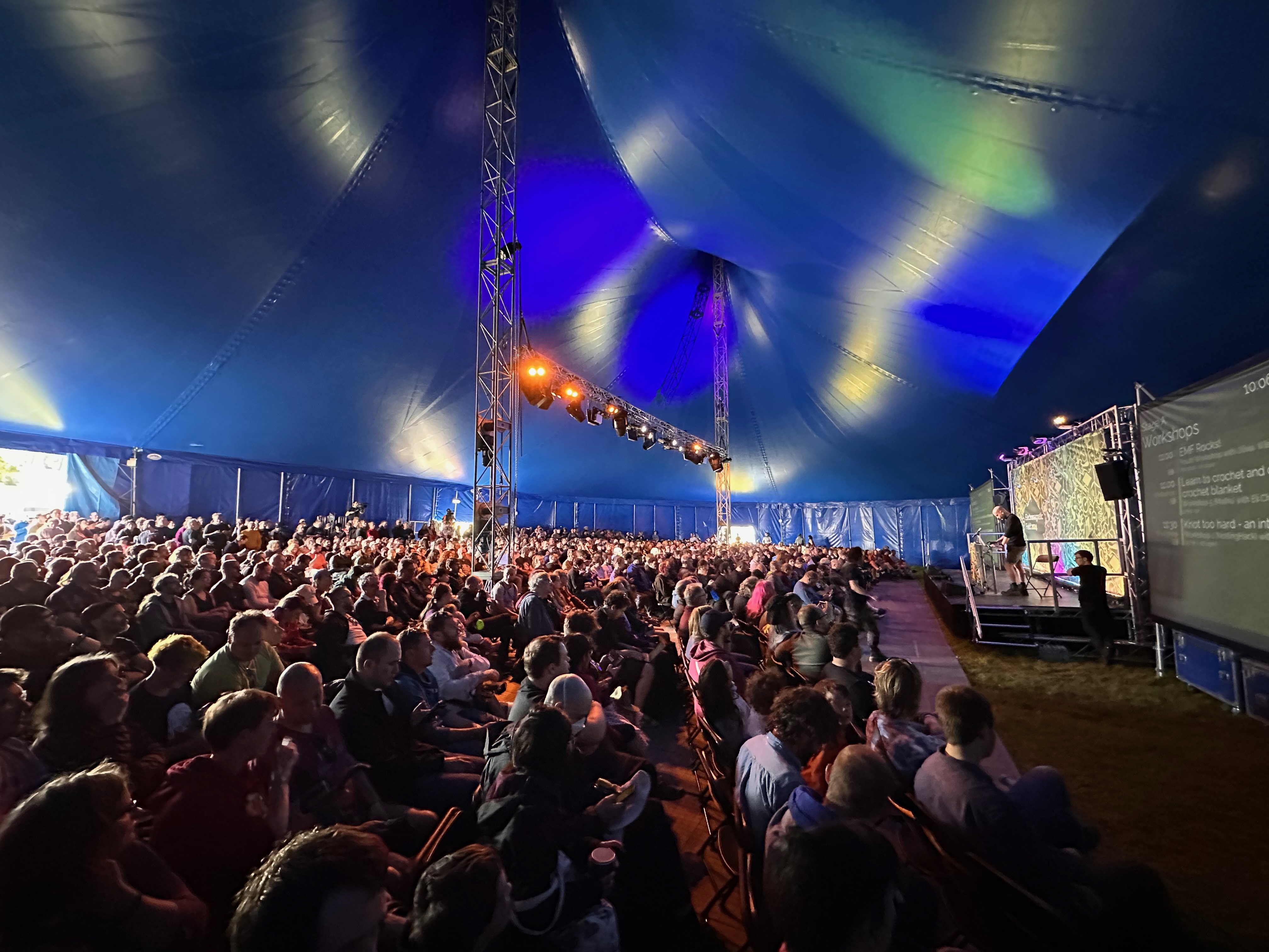 Photo of the opening ceremony at EMF. Looking from right to left across the audience. The circus style tent is packed, lights shine towards a single speaker on stage.