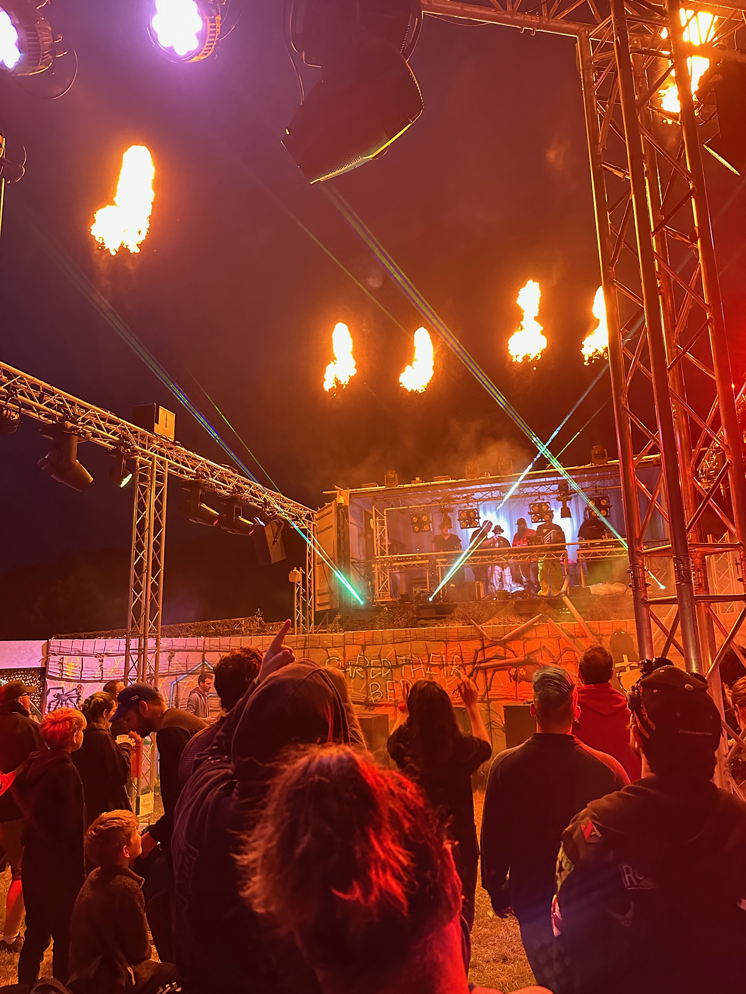 A photo looking over the dance floor. A raised DJ booth stands at the front of the dance floor, green lasers projected from its base. Behind the perimiter fire torches light the scene.
