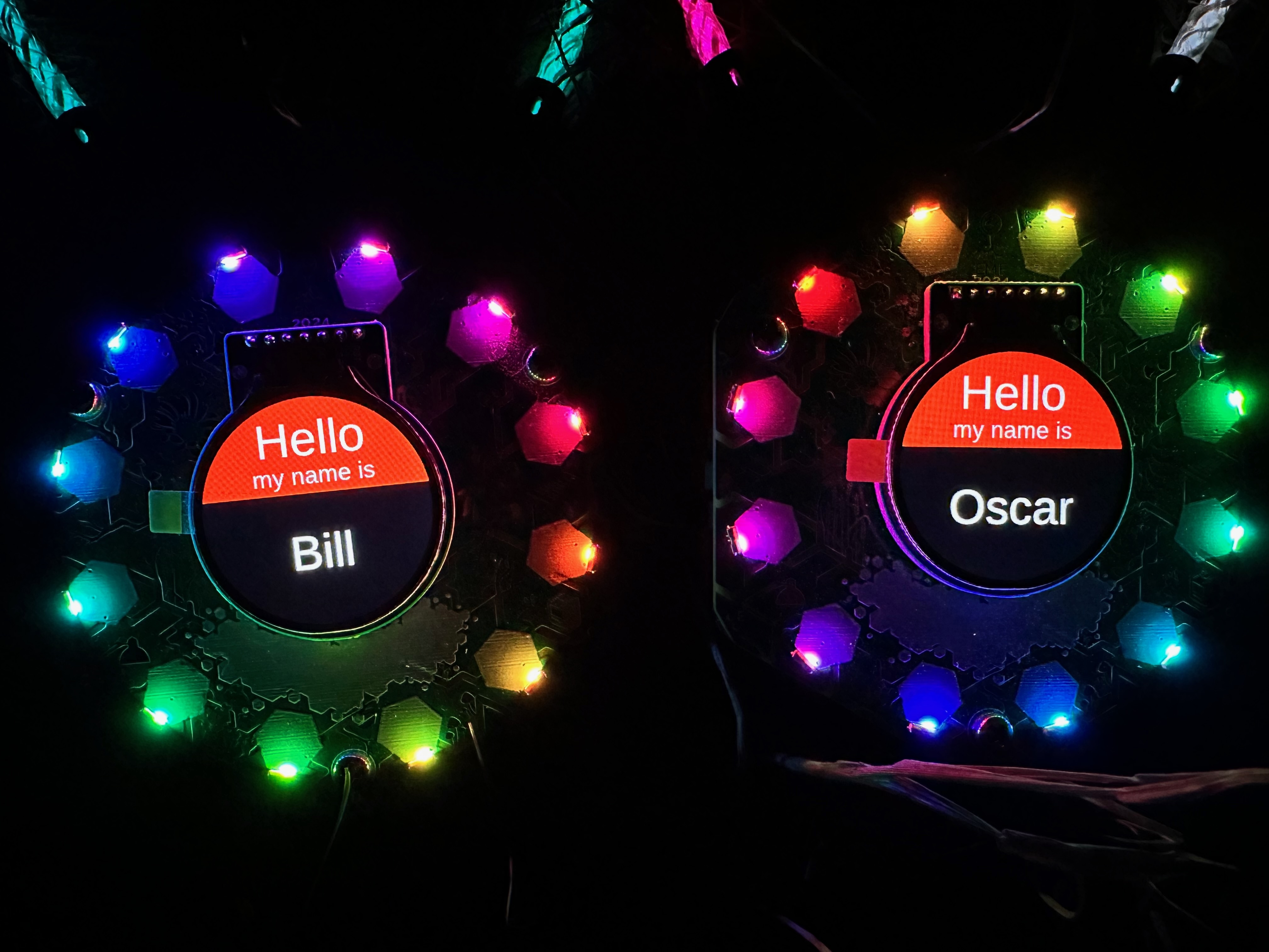 Two Tildagon badges surrounded by rainbow LEDs. In the centre of each is a message. 'My name is'... the left badge reads 'Bill' the right badge reads 'Oscar'.