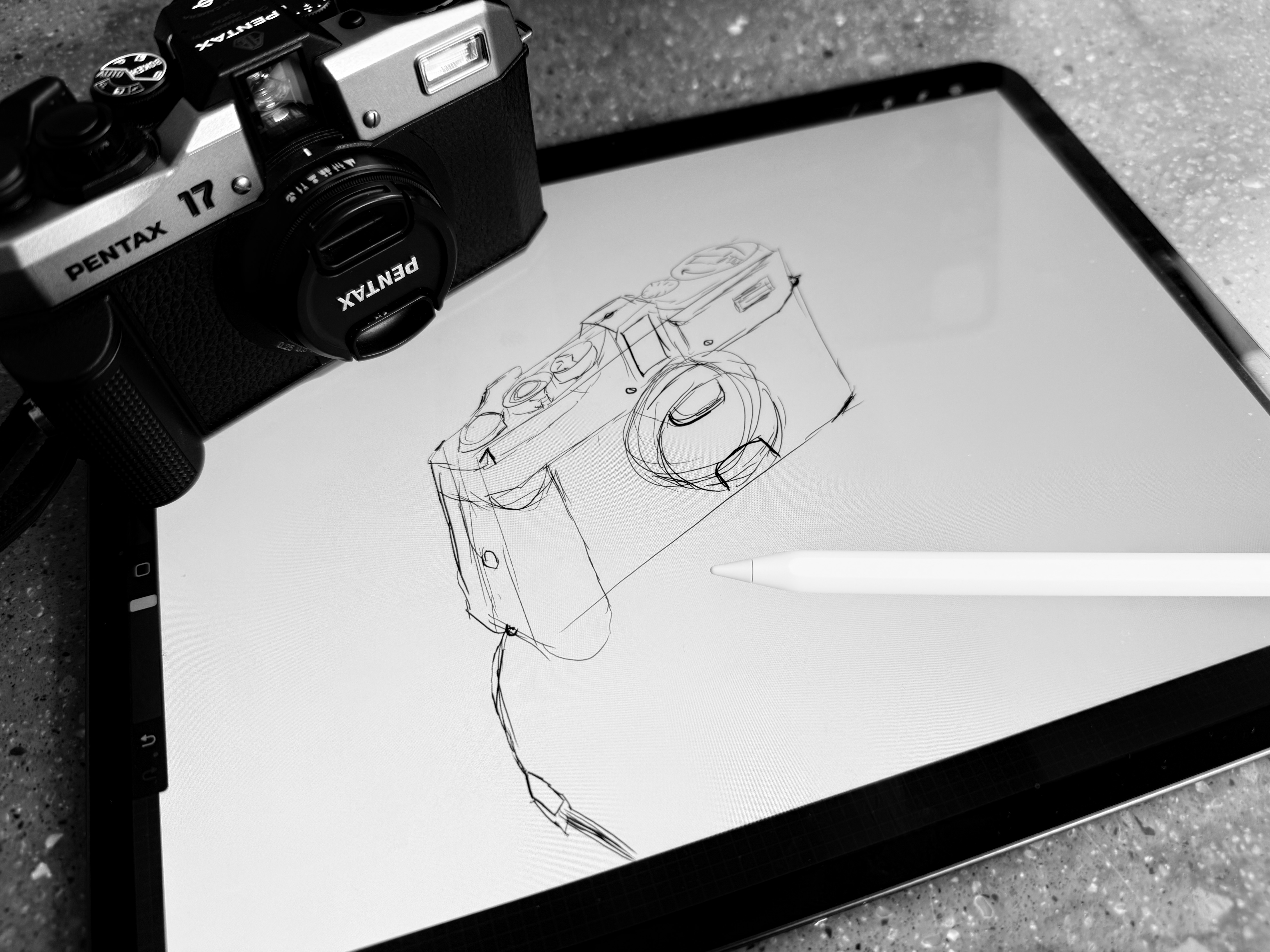 A black and white photo of an iPad laying on a kitchen worktop. An Apple pencil rests on the screen of the iPad. The image shown on the screen is a line sketch of a camera. On the top left corner of the iPad sits a Pentax 17 camera facing towards the frame. The lens cap is still on.