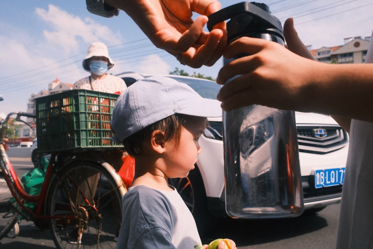 A boy looking out into the road. In his hand he is holding something. Behind him a car and a bicycle, in the foreground a water bottle