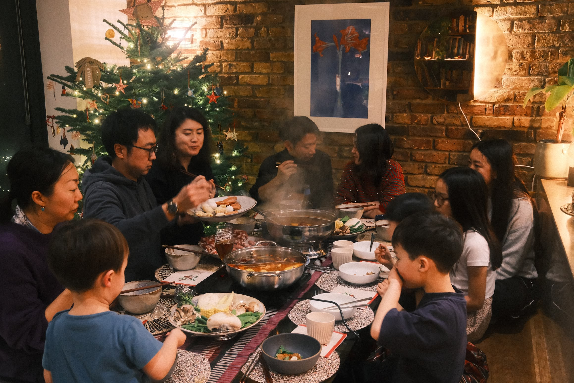 Families sit around a table covered in bowls of food, steam rising from cooking pots. In the background a Christmas tree lit with fairy lights