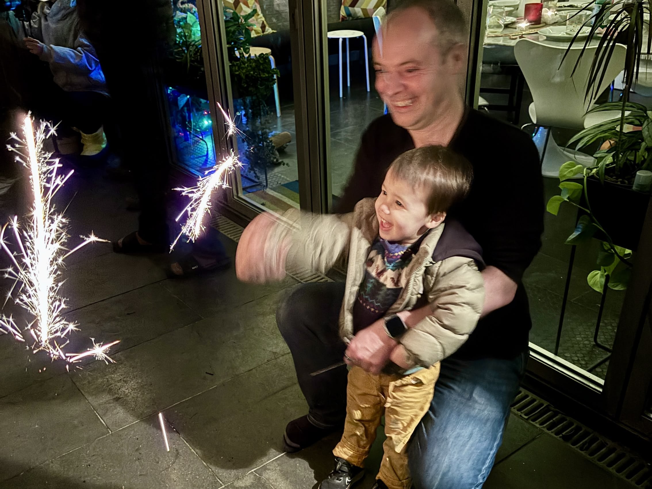 A boy holds a sparkler, sheer delight on his face, behind him a man kneels with his arm around the boy's waist