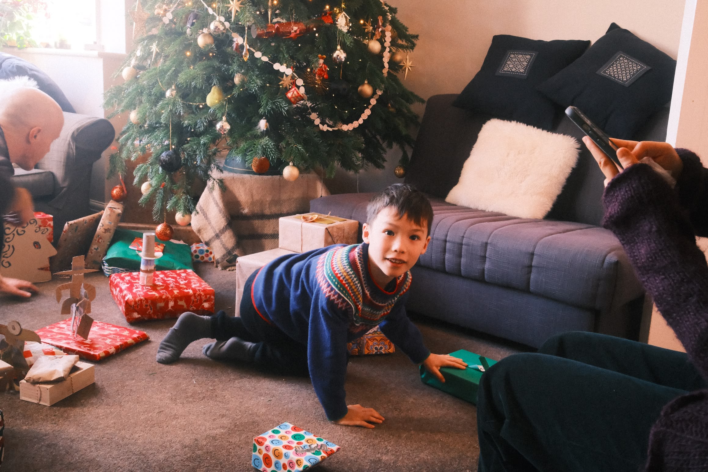 A boy kneels on the floor in front of a Christmas tree. Around him, a number of wrapped presents