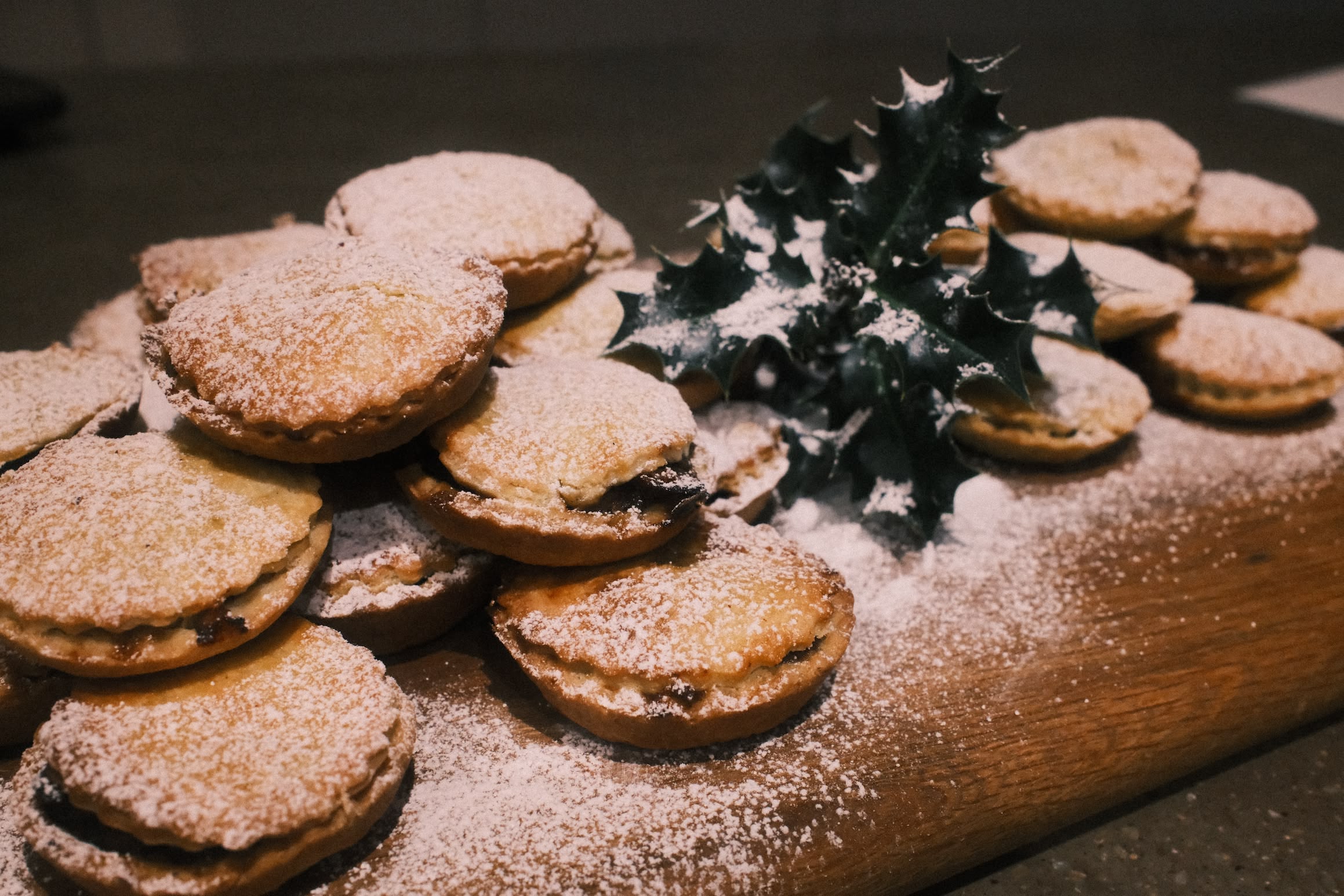 A pile of mince pies on a wooden board, a sprig of holly sits on the pile, a dusting of icing sugar gives it a wintery feel