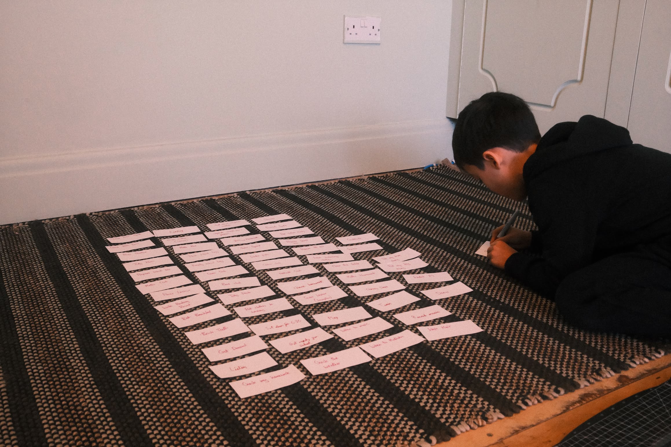 A boy kneels on the floor next to a grid of paper slips. He is writing something on one of the slips.
