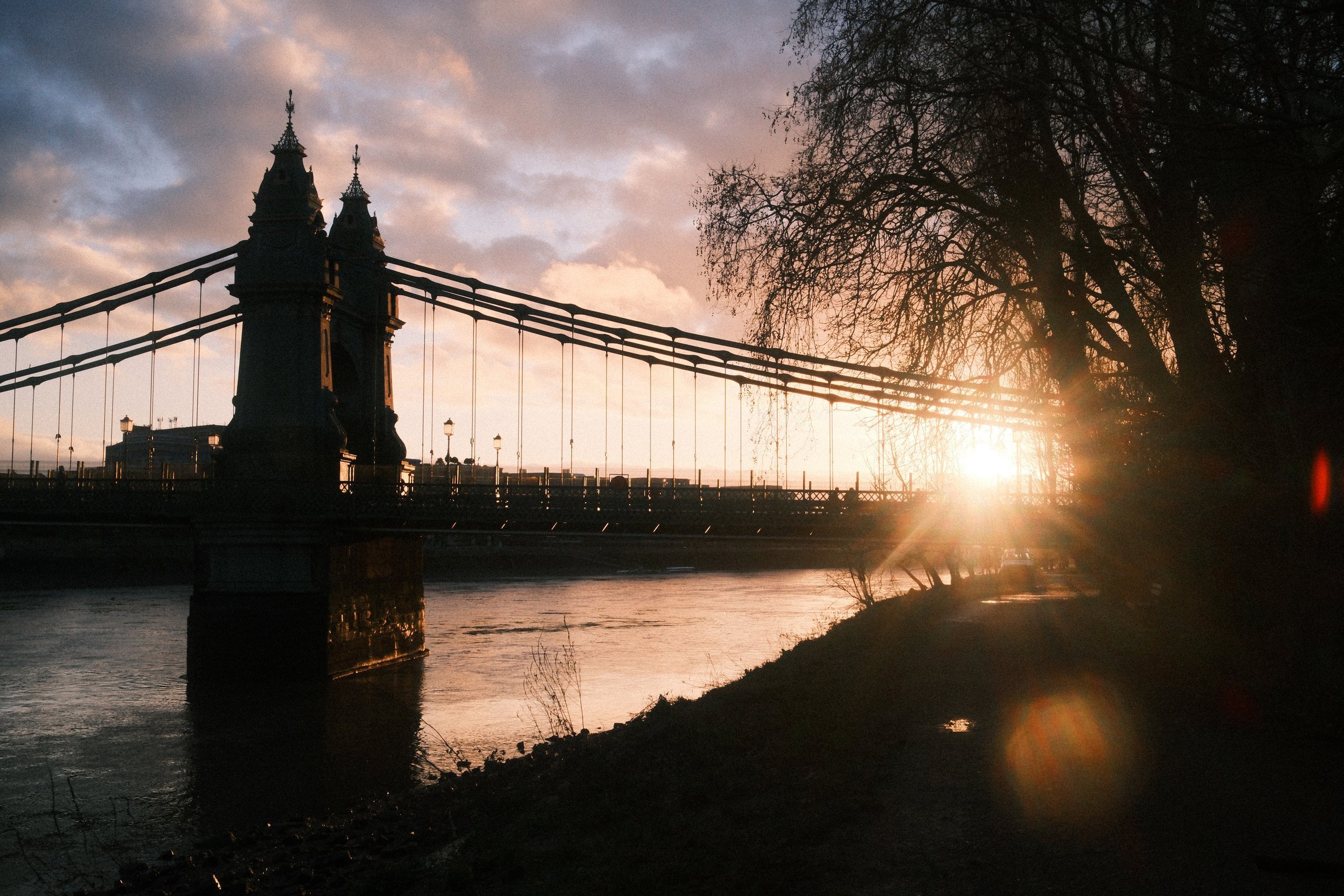 a photo taken into the sun looking along a river, a bridge silhoutted against the sunrise, trees hang over the near bank of the river
