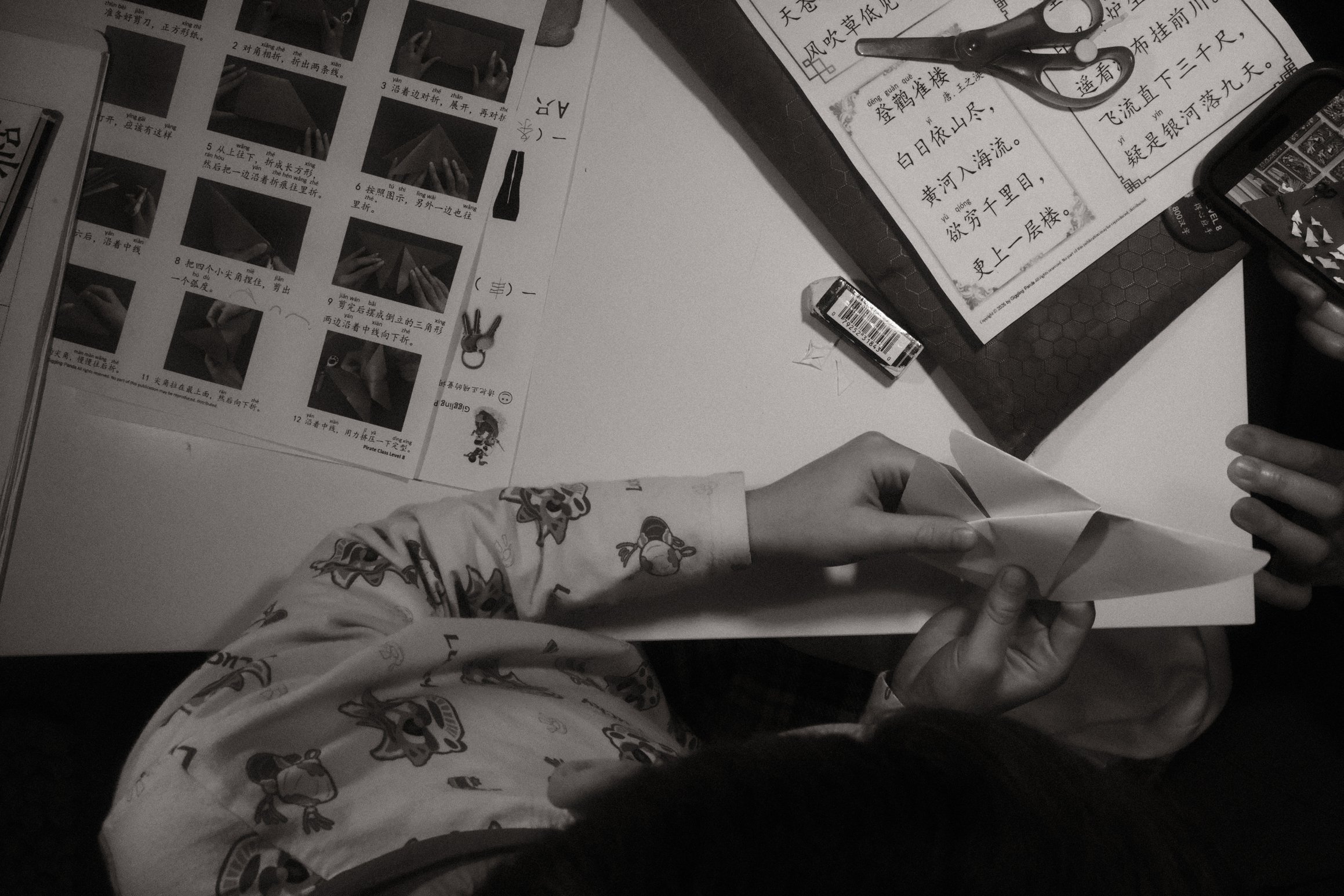 A black and white photo looking down on a child's desk. The child is folding origami. On the desk are a series of instructions in Chinese.