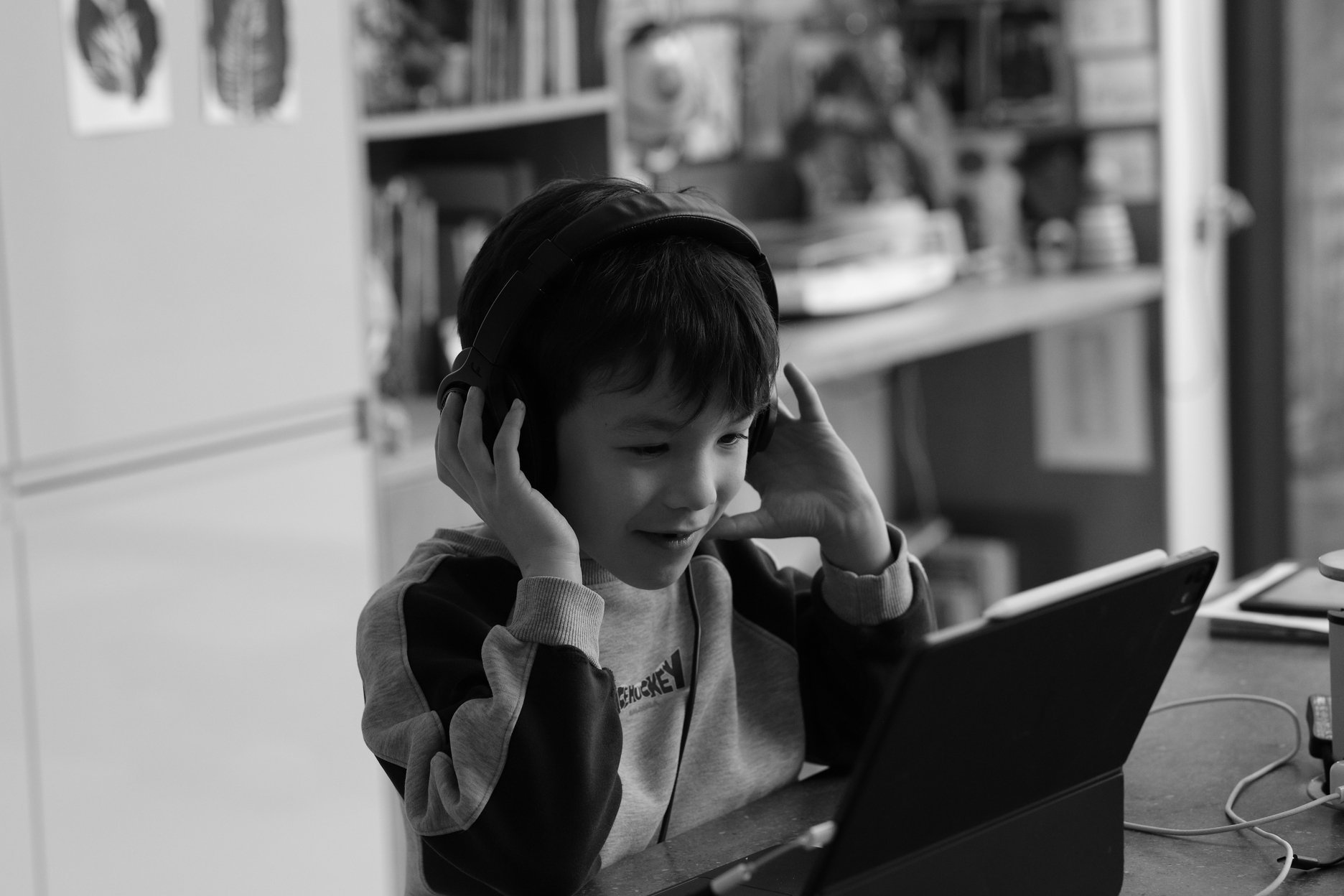 A black and white photo of a boy wearing headphones smiling at an iPad. His hands holding the cups of the headphones as if he's about to take them off.