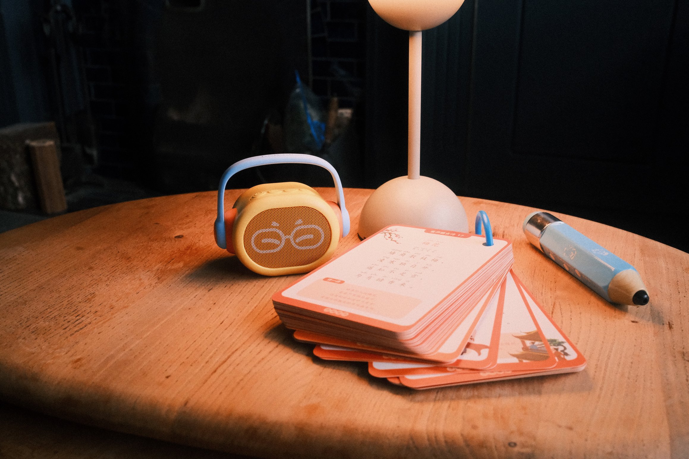 A wooden table. On the table sit a small speaker with a handle, a deck of cards on a ring binder, a large pen, and a lamp