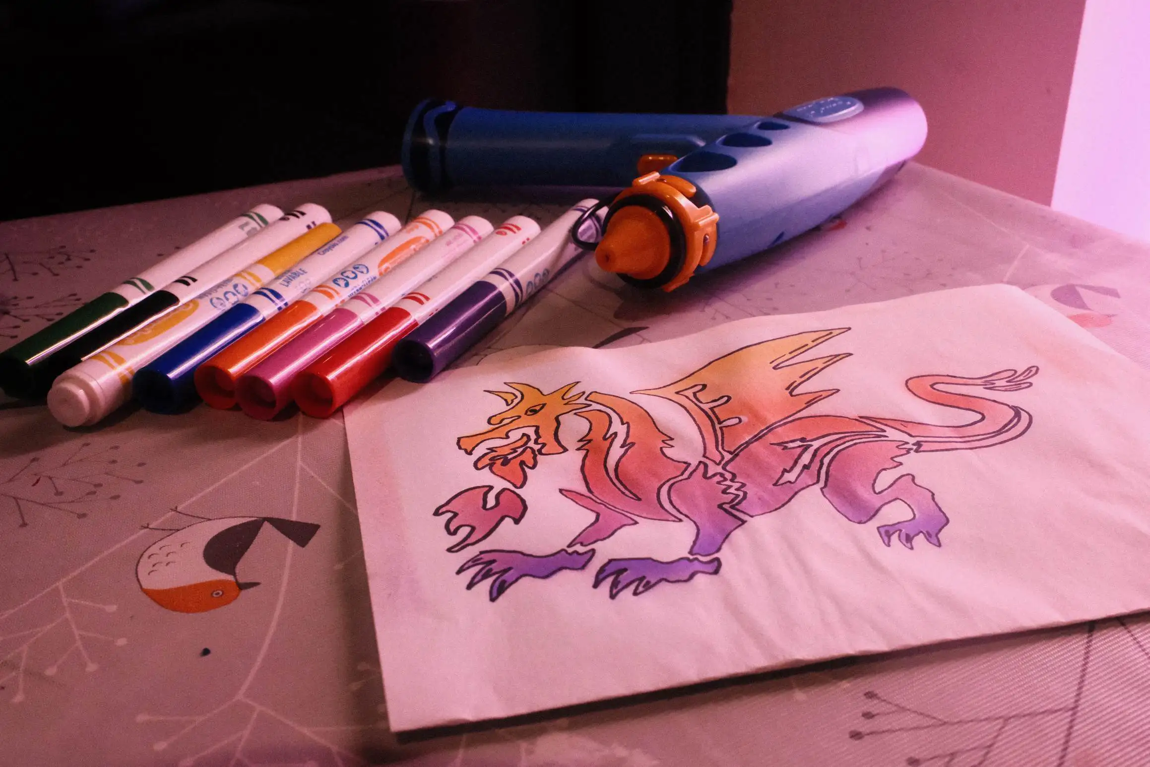 An image of a dragon on a folded sheet of white paper. The bottom half of the dragon is in purple, moving through red and on to yellow as you move up the dragon. In the background there are an array of watercolour pens and a kids airbrush.