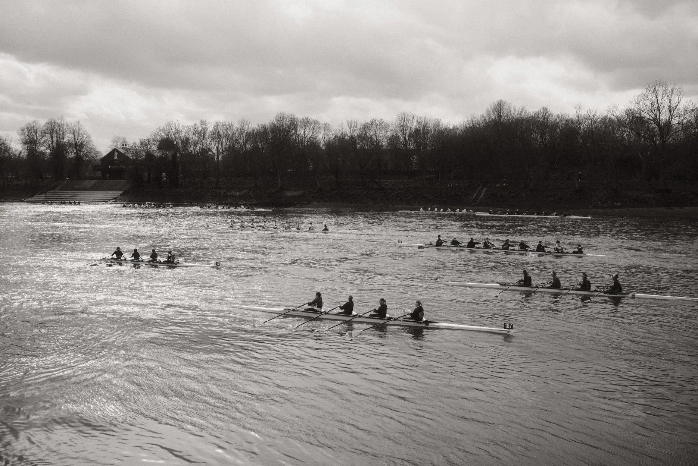 A black and white photo of a river. 9 crews jostle for position before the start of the race. The quads are closer to the bottom of the frame, the eights line up on the far side of the river.