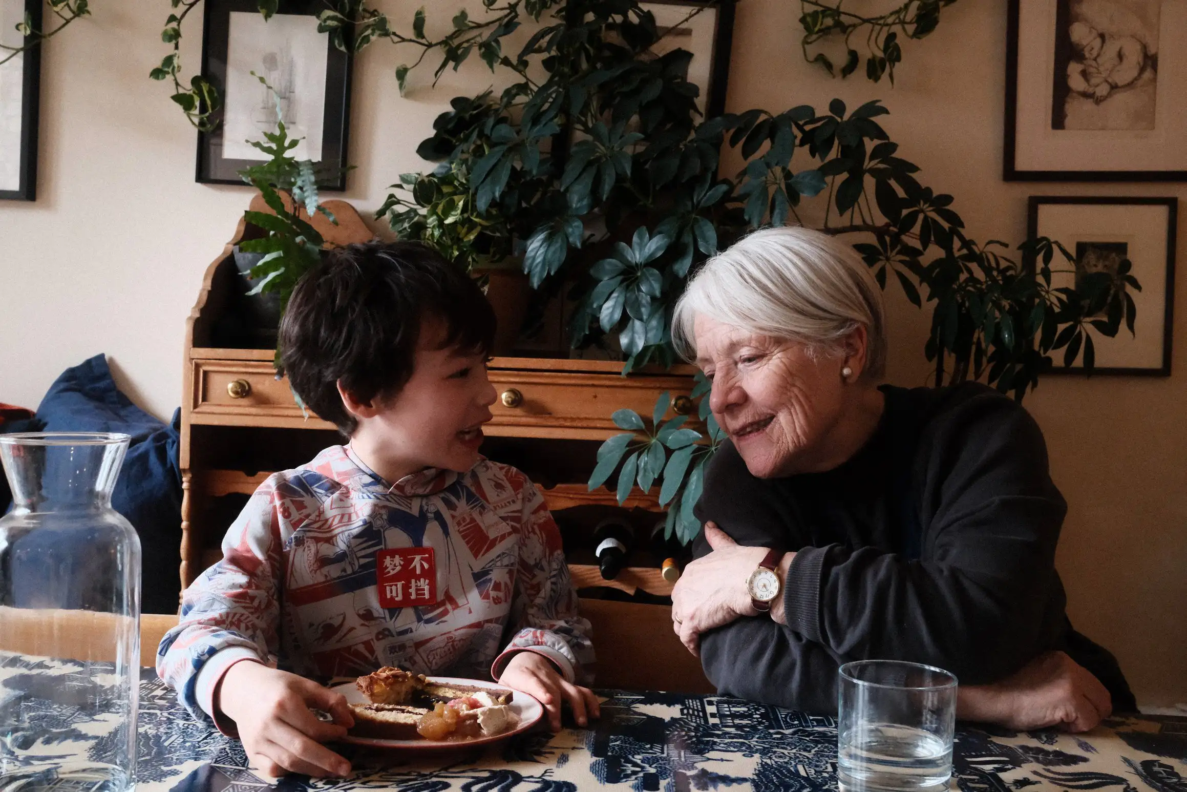 A boy and his grandmother engaged in conversation. In front of the boy is a plate of various deserts.