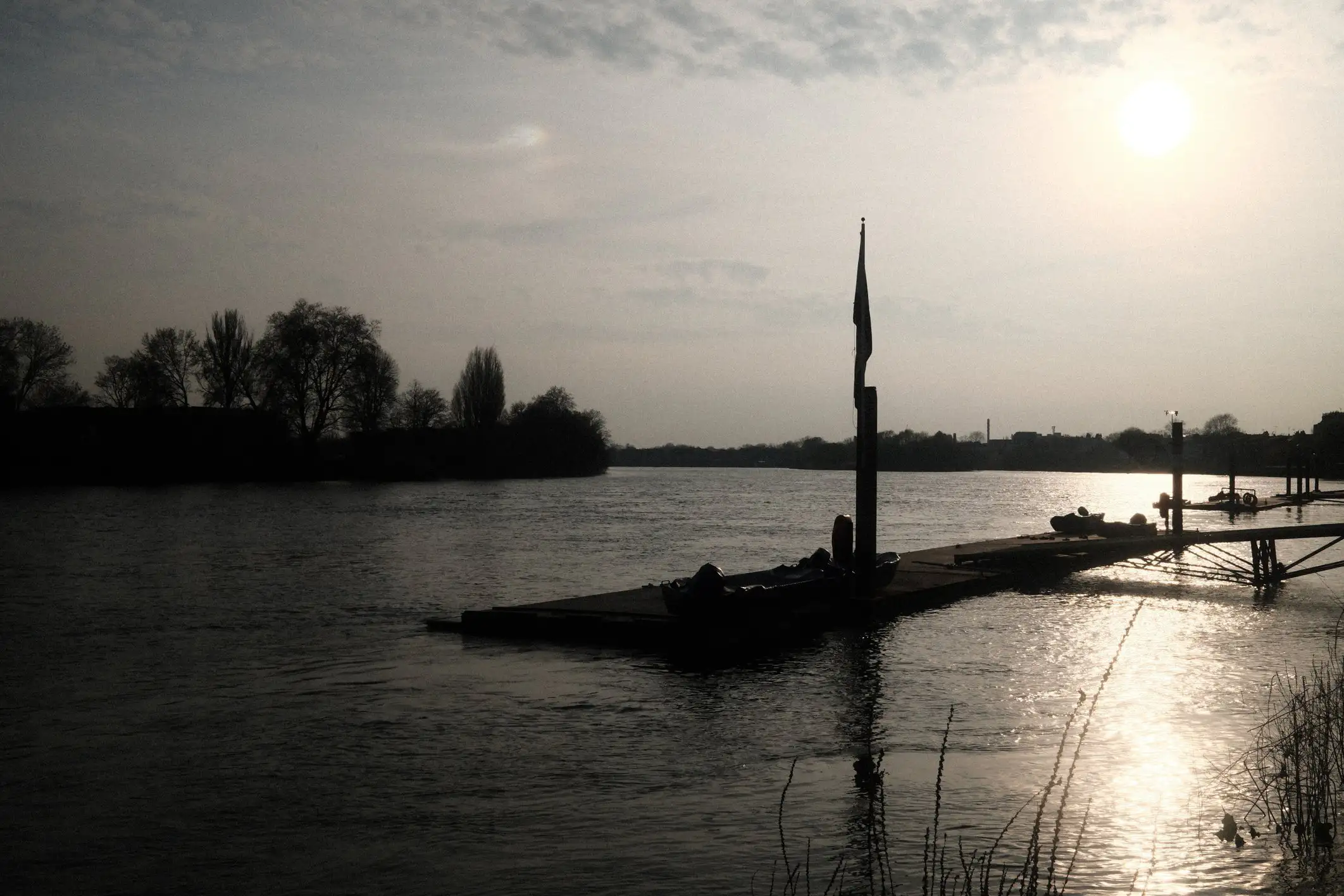 A photo looking West along the Thames, the sun setting in the distance, a jetty silhouetted against the cool evening sun