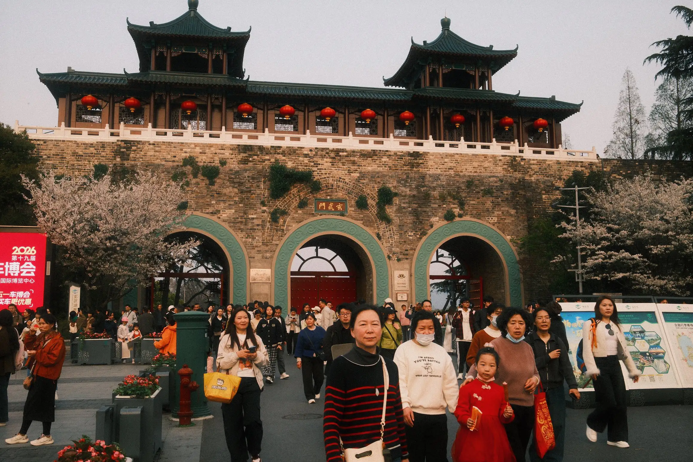A crowd of people leave the gate to the Xuanwu Lake in Nanjing. At the front of the crowd, a family, the young daughter dressed in red, look towards the camera.