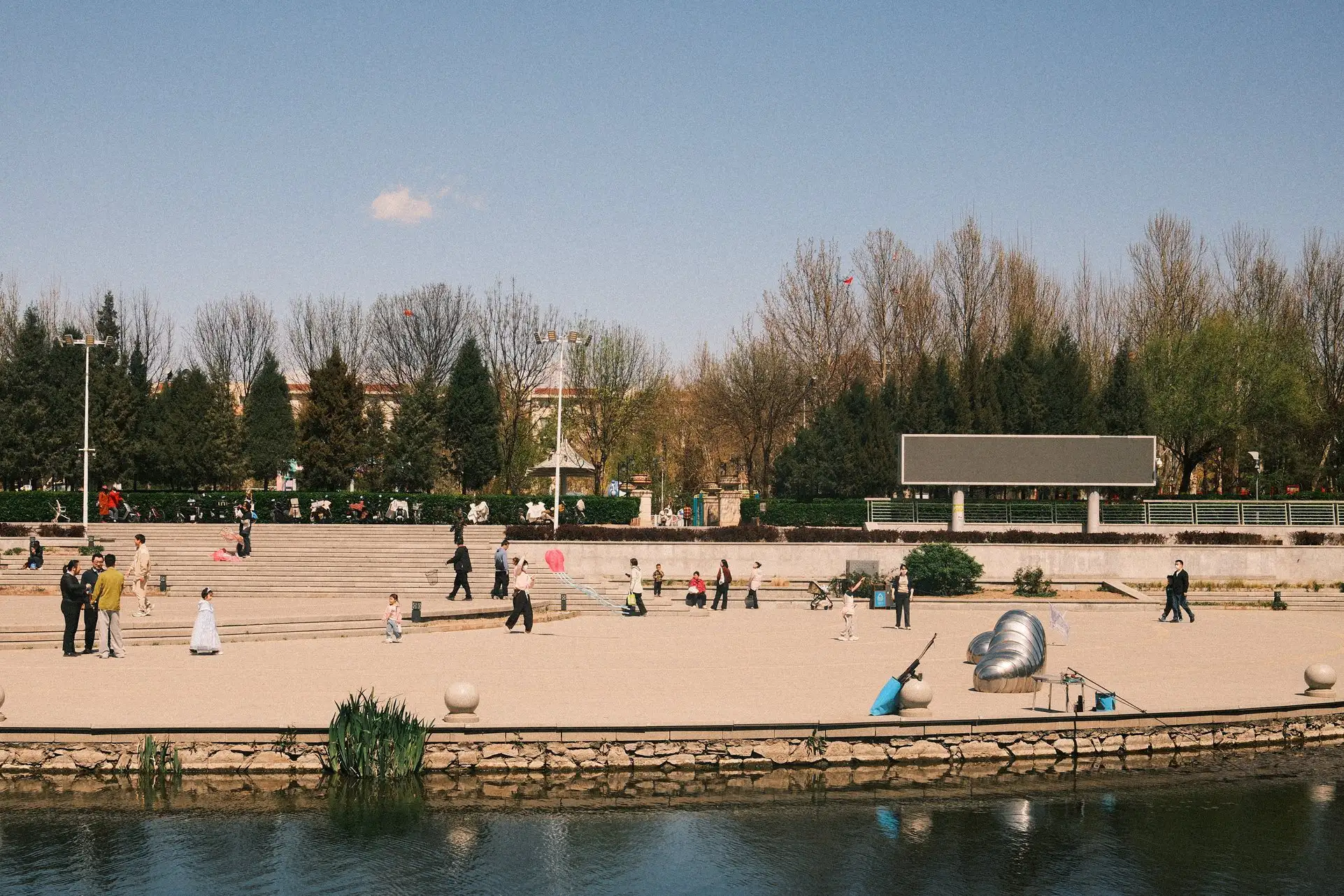 A photo looking over a small river to a paved area on the opposite bank. Families can be seen enjoying the sun; talking, fishing, flying kites, holding balloons.