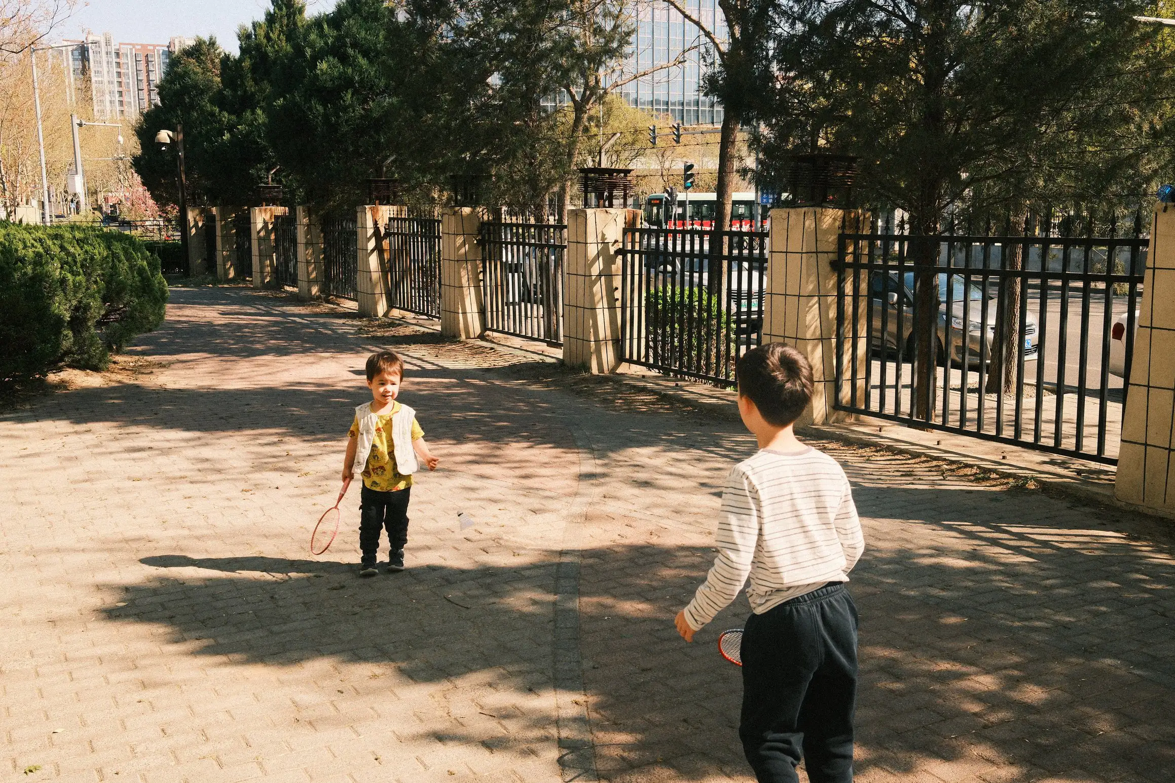 Two boys play badminton together on a paved area fenced off from a road.