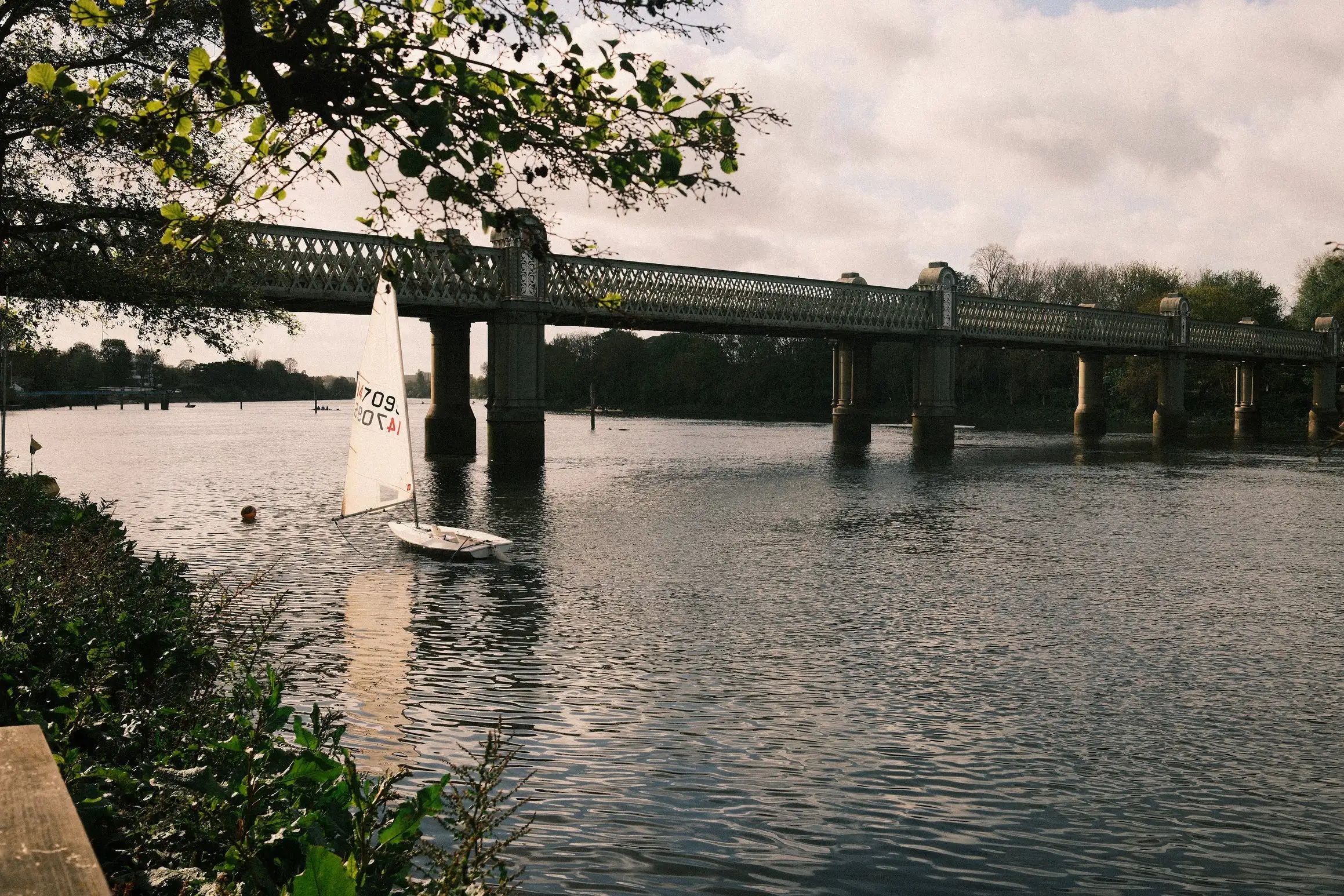 A photo looking along a river towards a railway bridge. A small sailing boat sits on the water. No one is on the boat.