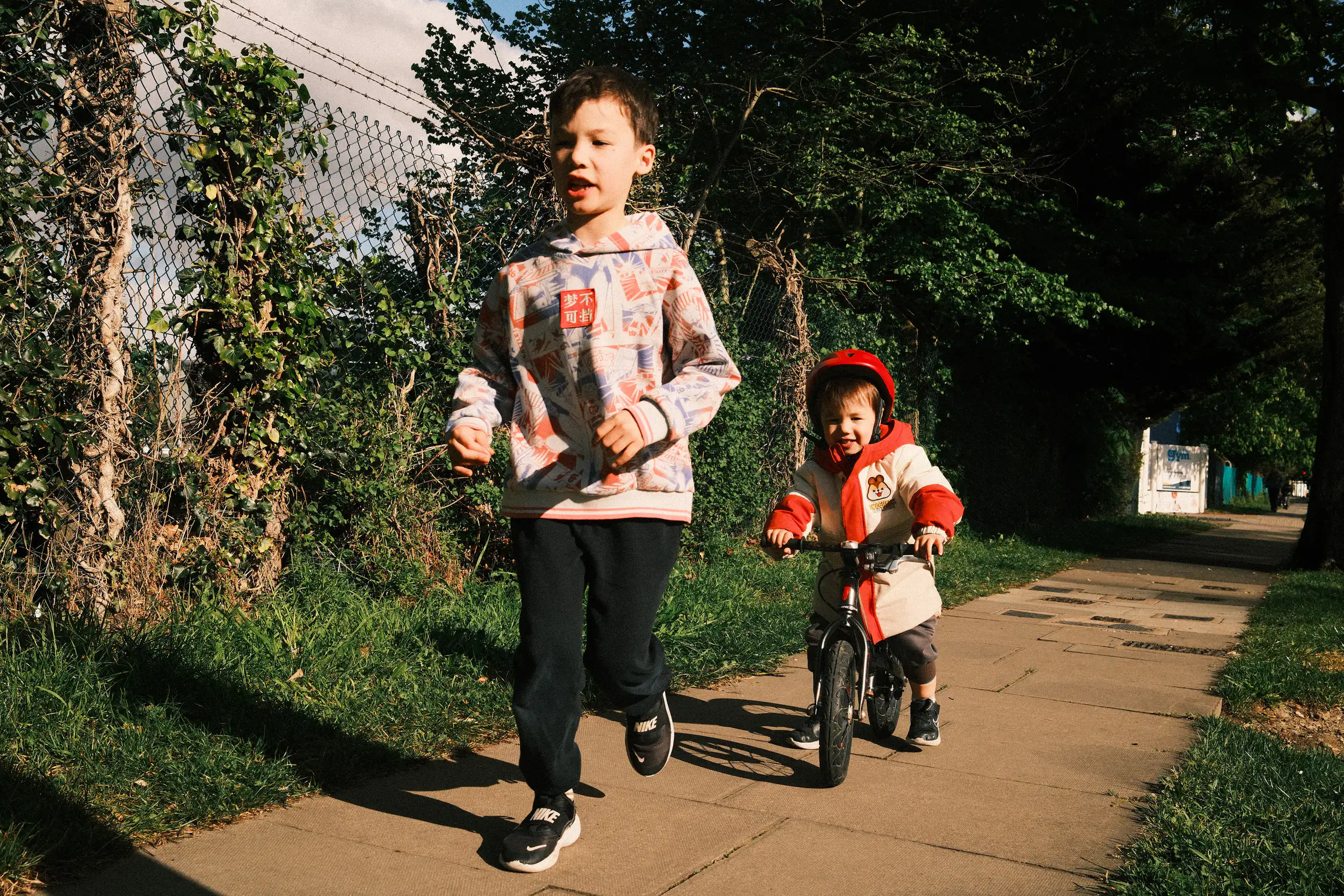 Two boys head along a footpath, the elder one running, the younger on a balance bike.