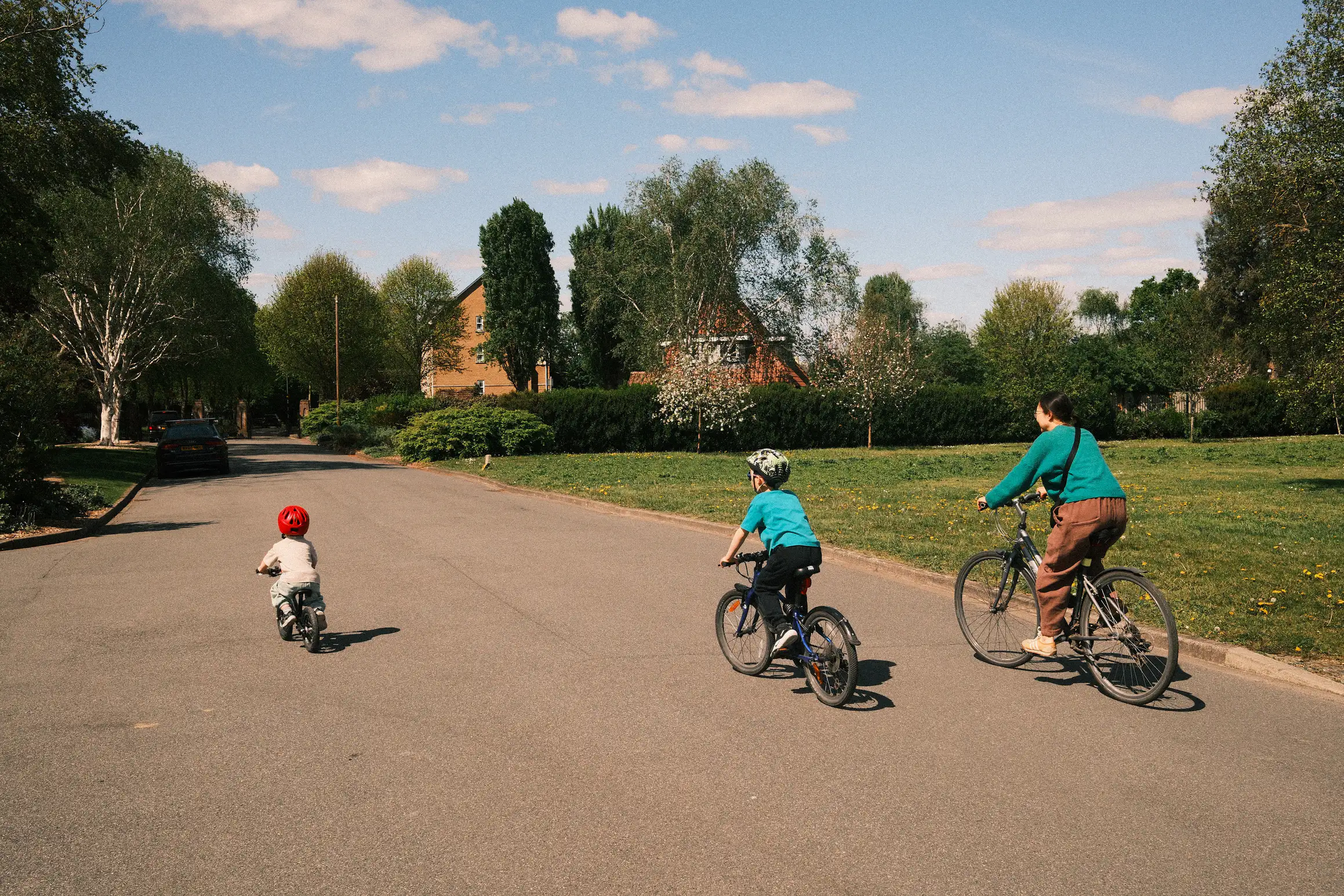 two young boys and their mother cycle along a quiet road, the youngest is in front on a balance bike, feet off the floor