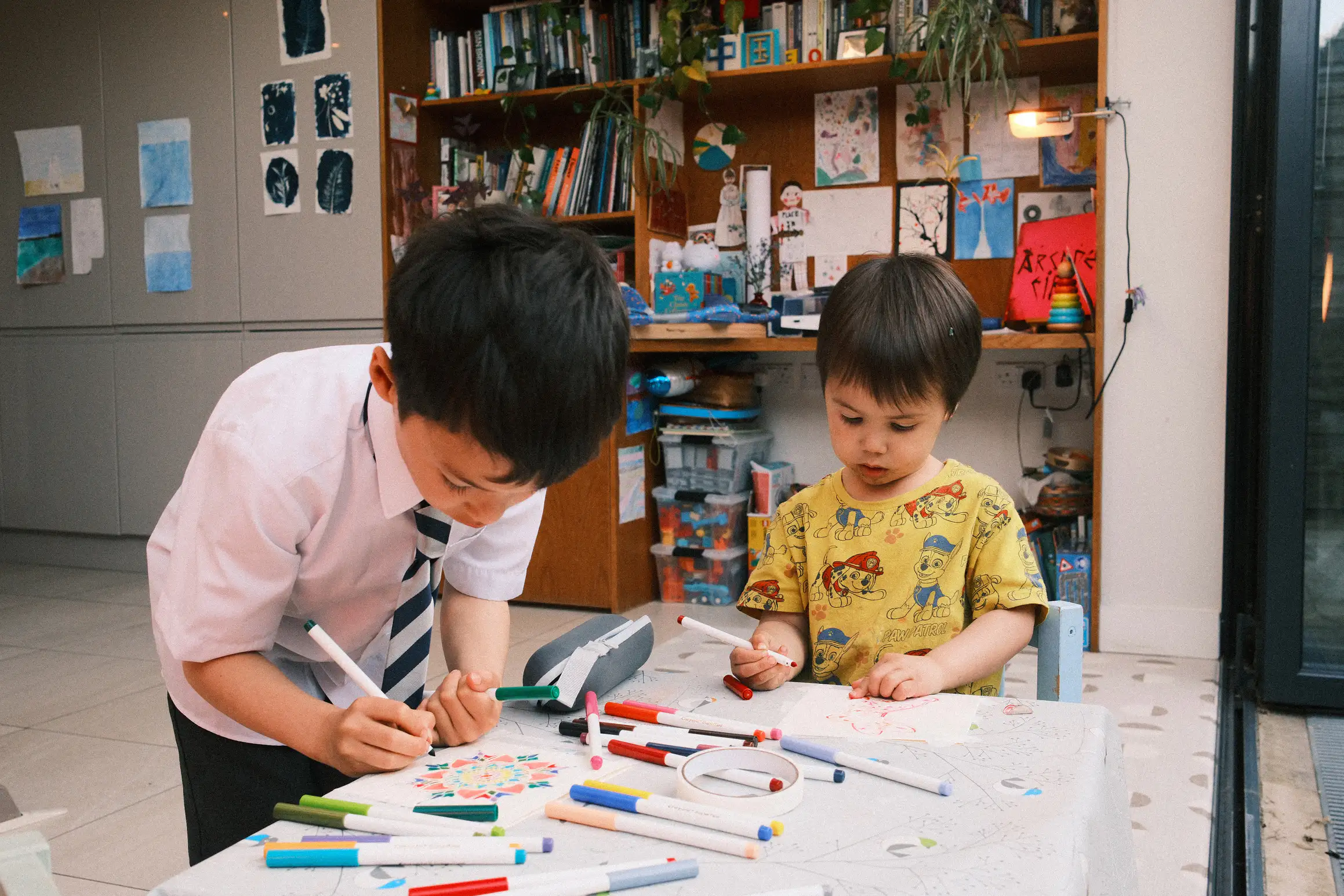 two boys hunched over a small table making greetings cards, the table covered in felt pens