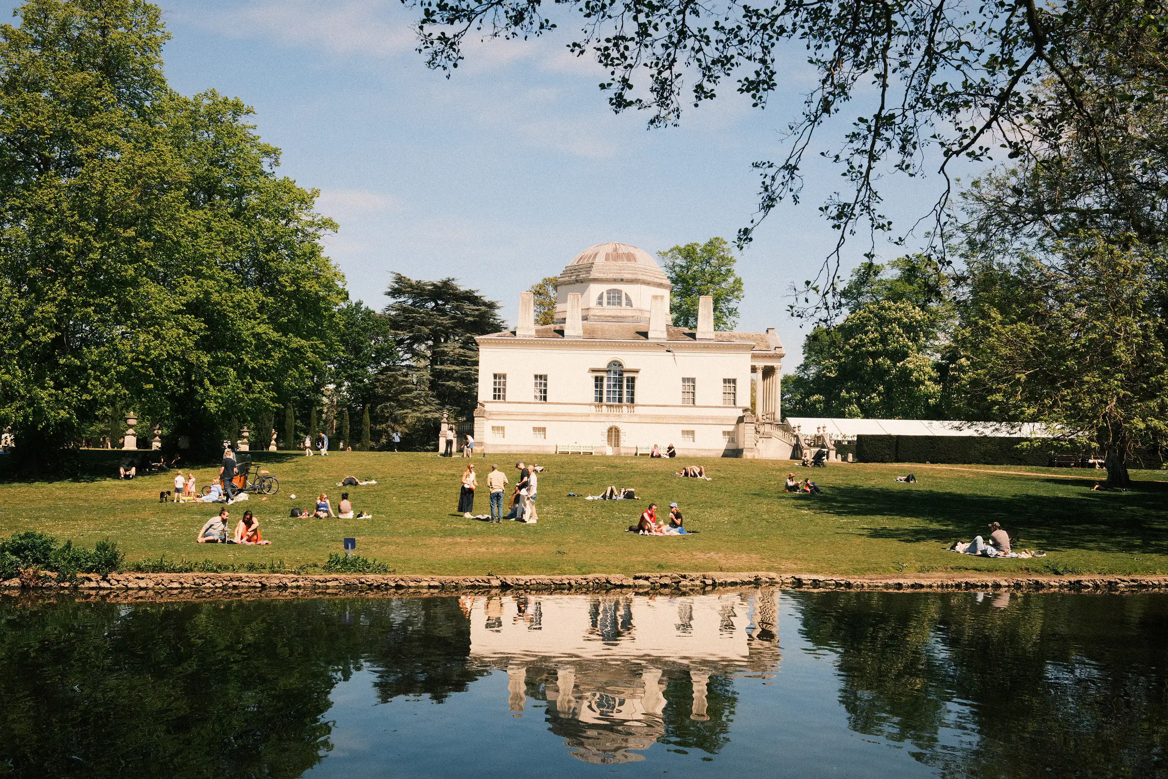 a view over the lake towards Chiswick House, people sat on the lawn enjoying the sunshine