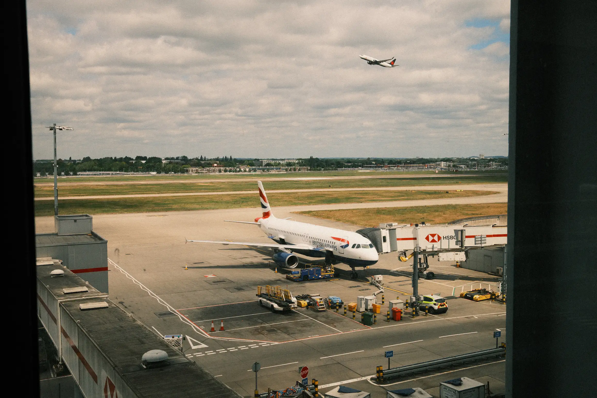 A British Airways plane sits on the apron at Heathrow. In the distance another flight can be seen taking off.