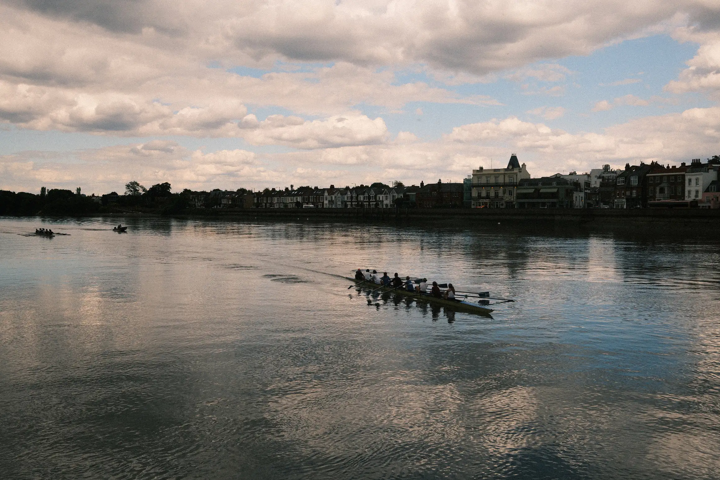 a crew of 8 row along the river Thames heading towards Barnes Bridge, on the far side of the river are the residential properties of Barnes