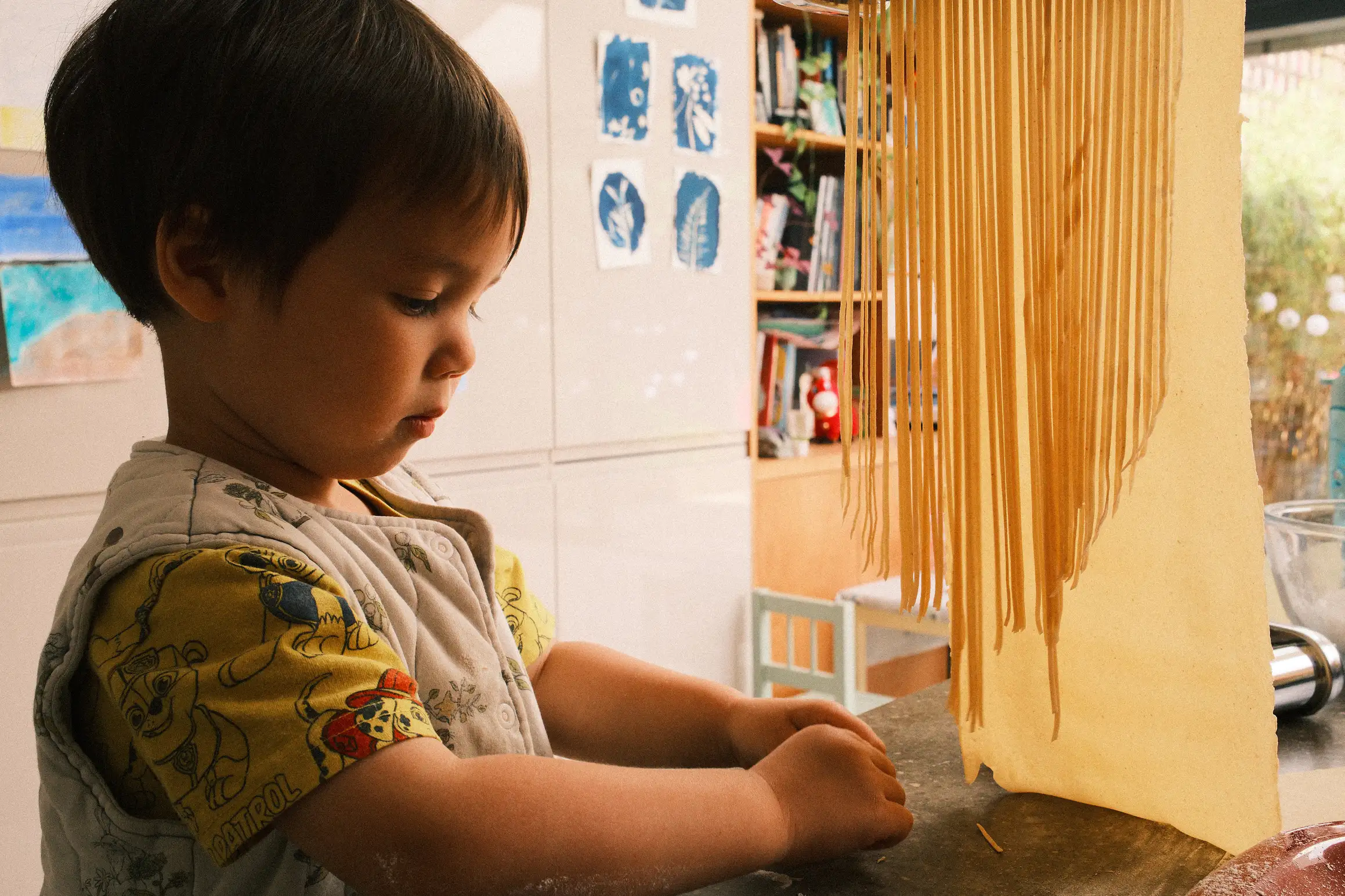 a boy gathers up pieces of dough that have fallen from a pasta machine, strands of pasta hang closes to the camera, a wide sheet of uncut pasta hangs behind them, out of shot the feed through a pasta cutter