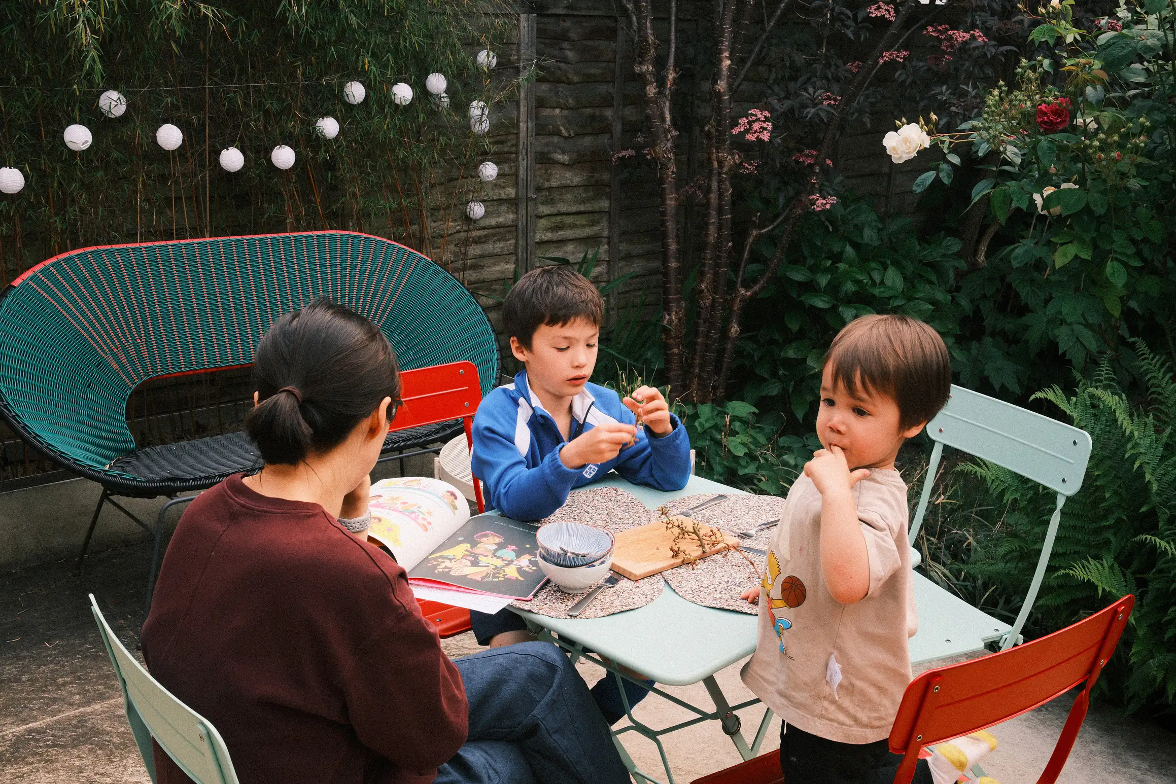 two boys and their mother sit at a table in the garden, empty dishes and grape stalks suggest they've just finished eating