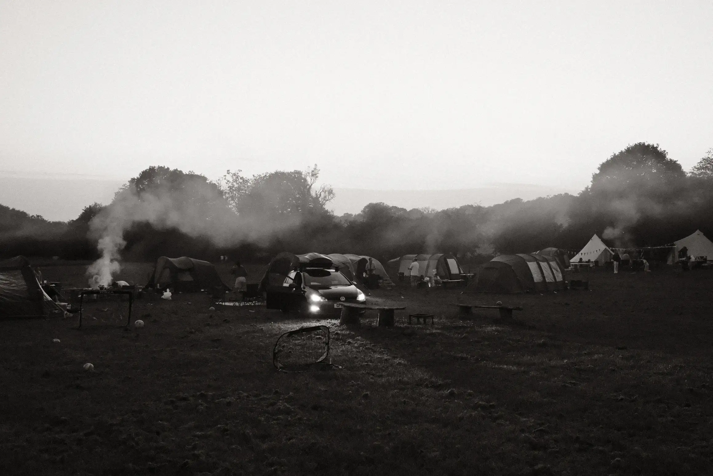 a black and white photo of a campsite, smoke rising from a couple of camp fires, a car with its headlights on faces towards the camera
