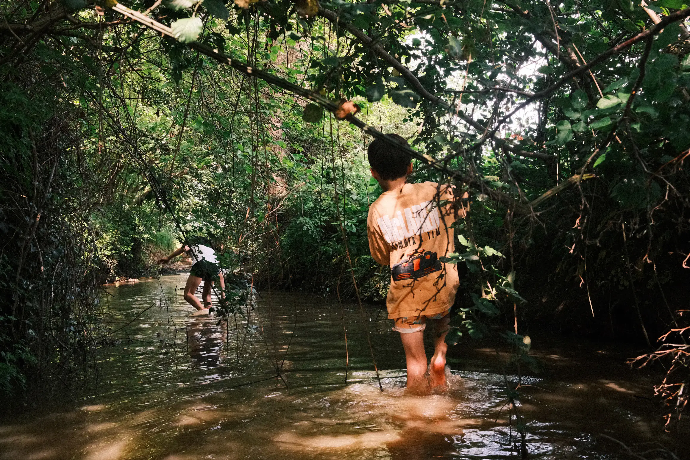 a boy follows a girl down a muddy stream, the water up to their ankles, either side of the stream branches and leaves provide shade from the sun