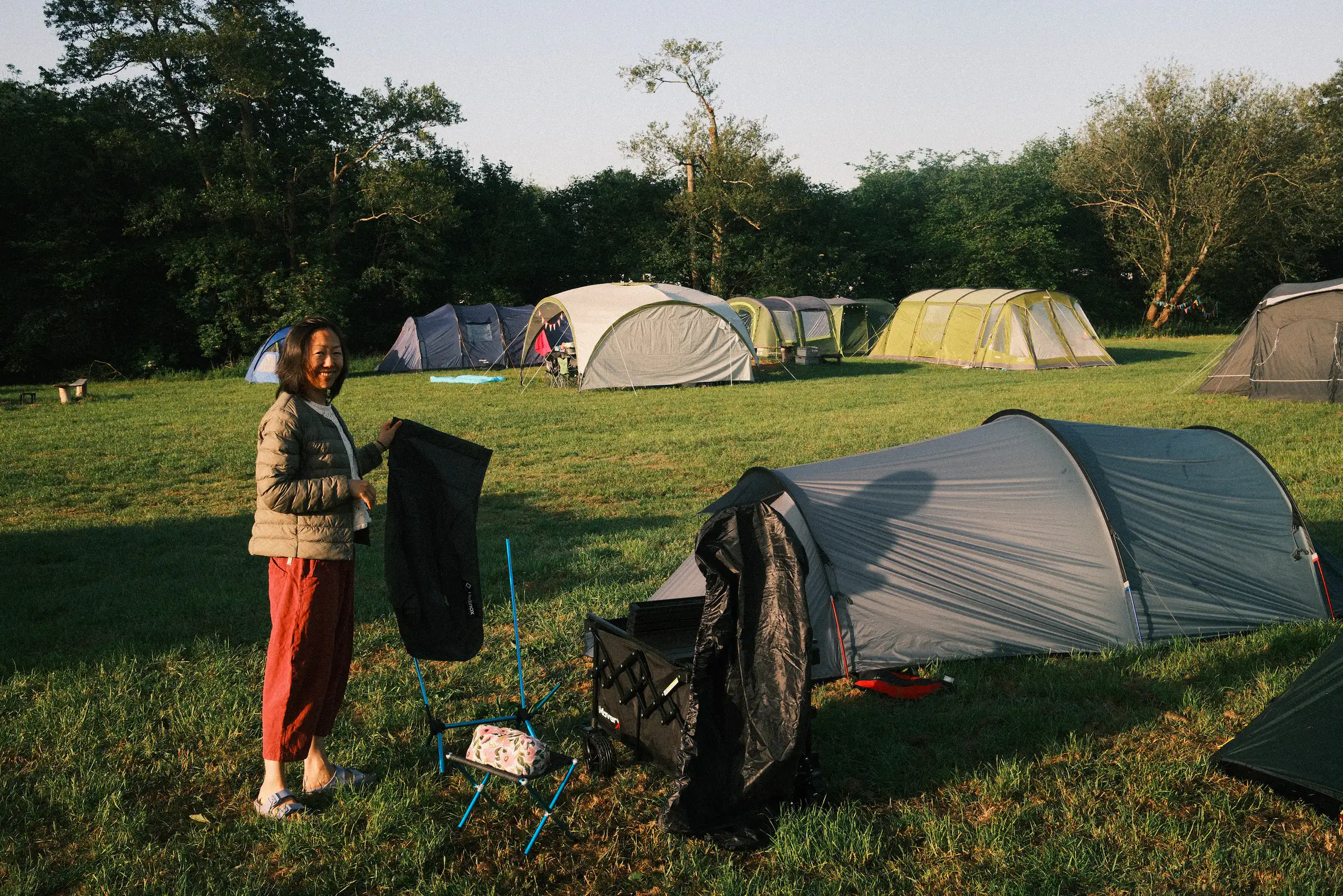 a person stands by a tent holding a bag, the frame of a chair next to them suggests they are either packing or unpacking the camp