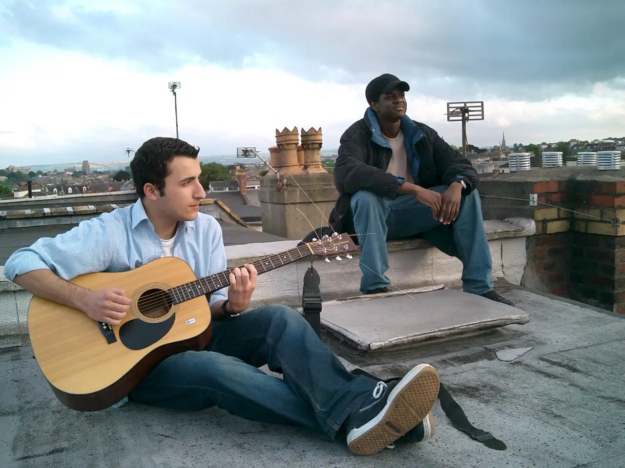 Two people sit on the roof of a house, one plays a guitar.