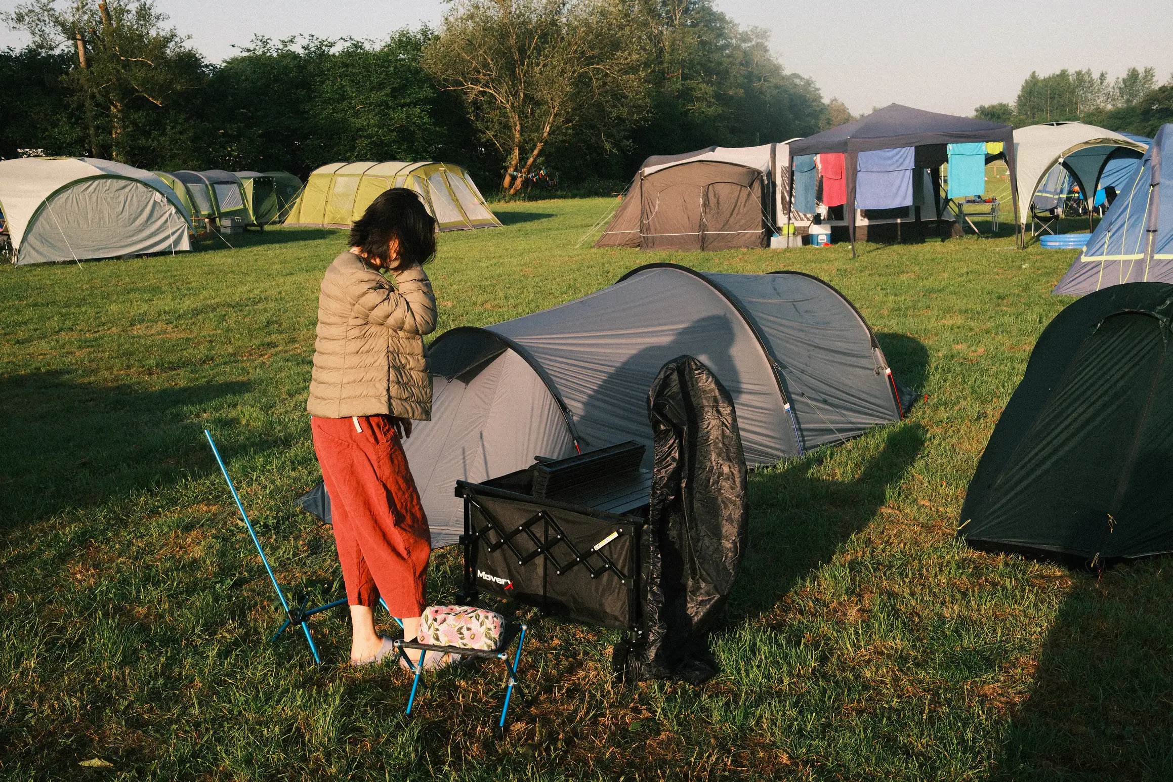 a person stands in a field looking at a camping trolley, a number of family size tents in the background, no other people are visible