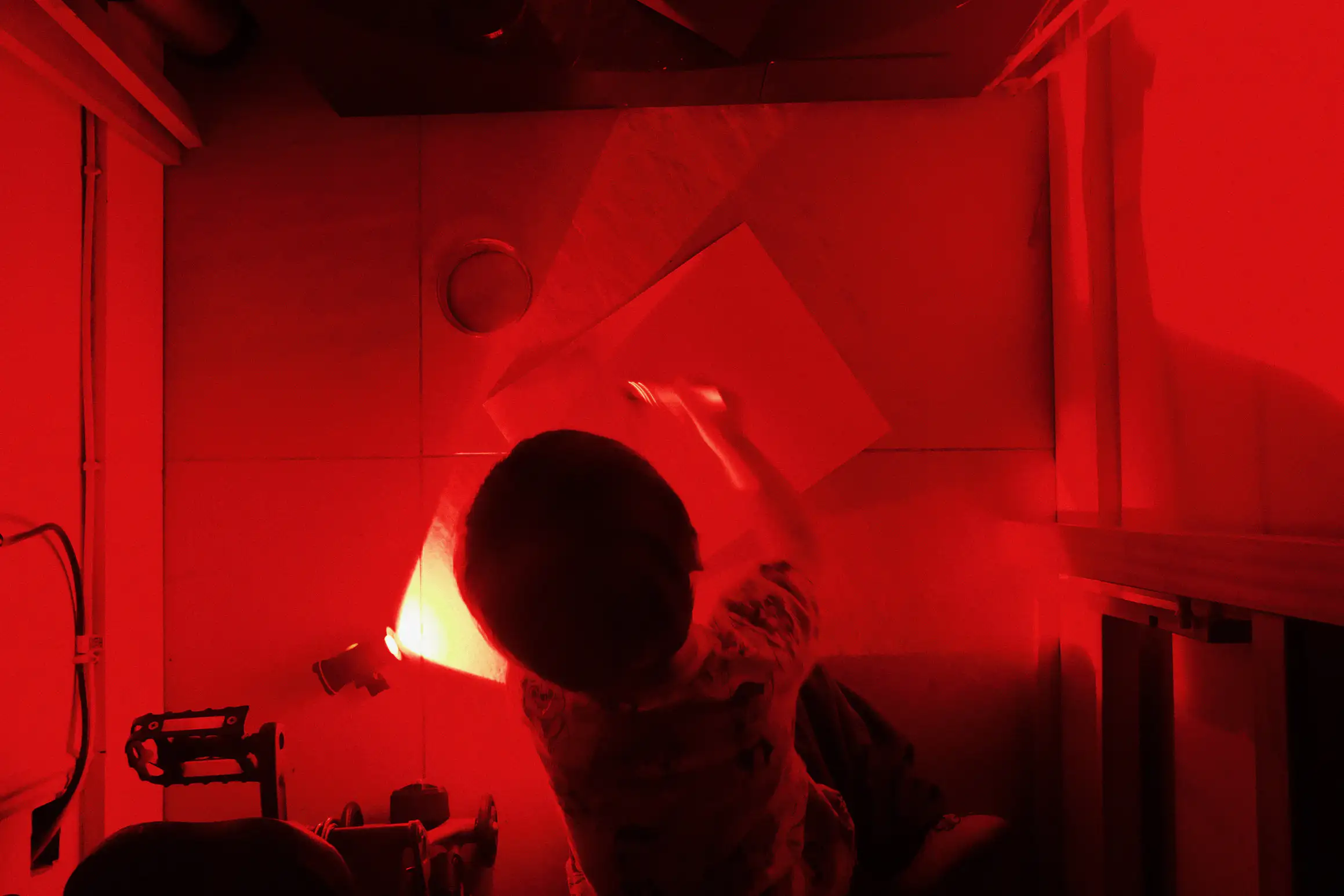a boy sits on the floor painting light sensitive solution onto a piece of paper, the image is lit by light from a red bike light
