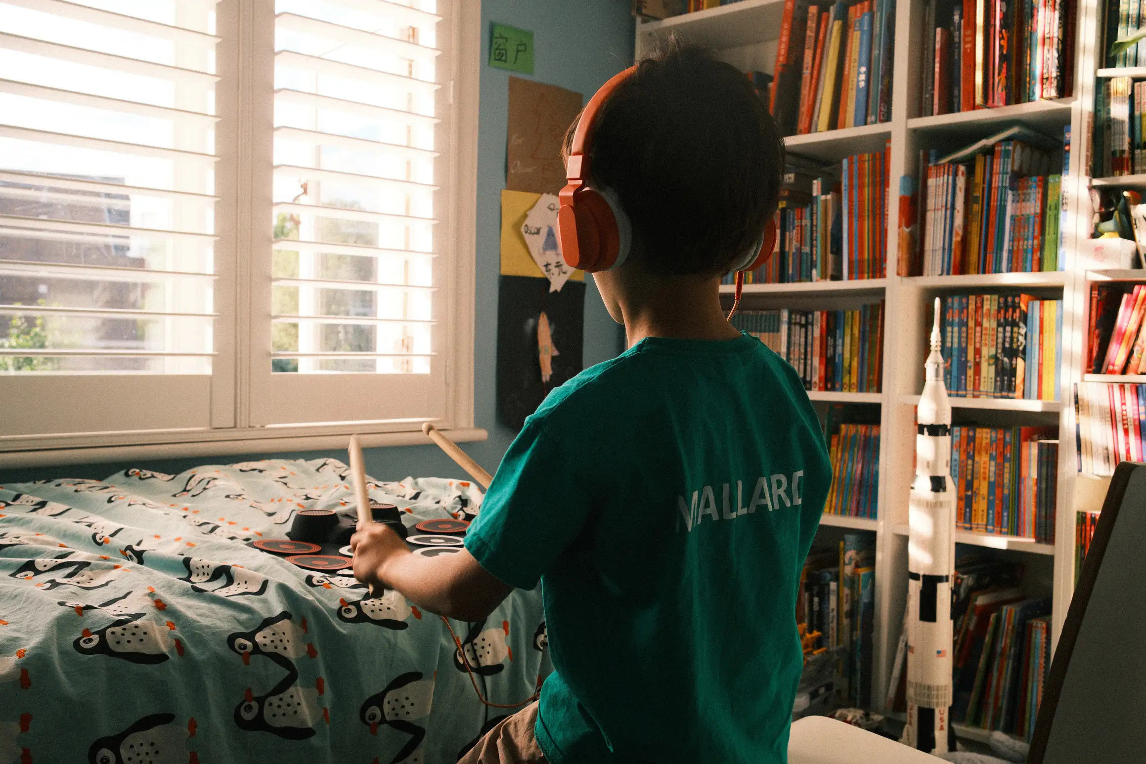 a photo of a boy playing a toy drum mat on his bed, behind him a bookshelf and the Lego saturn rocket