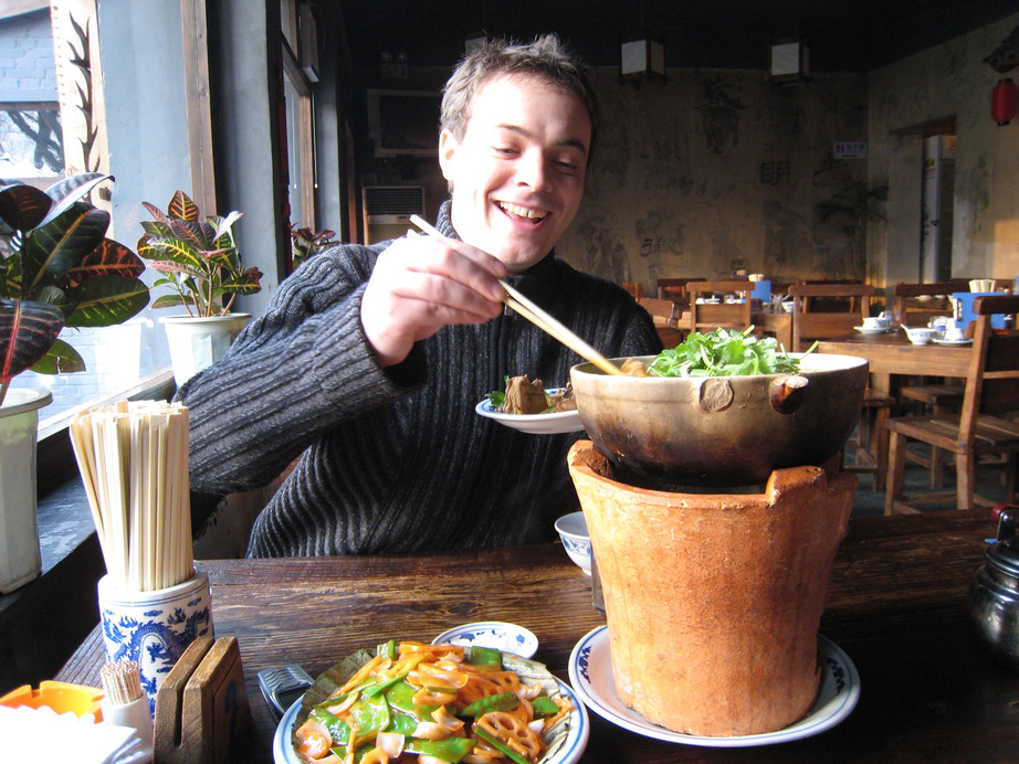a photo of a person sat at a restaurant table, a plate of stir-fried vegetables sits on the table next to a clay pot resting on a small stove. The person uses chopsticks to take food from the pot. The restaurant appears otherwise empty.