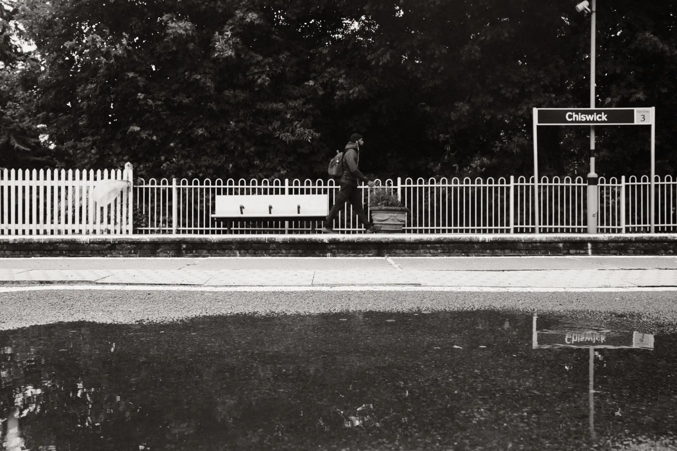 A black and white photo of a man walking along the station platform at Chiswick station. In the foreground a pool of water provides a reflection.