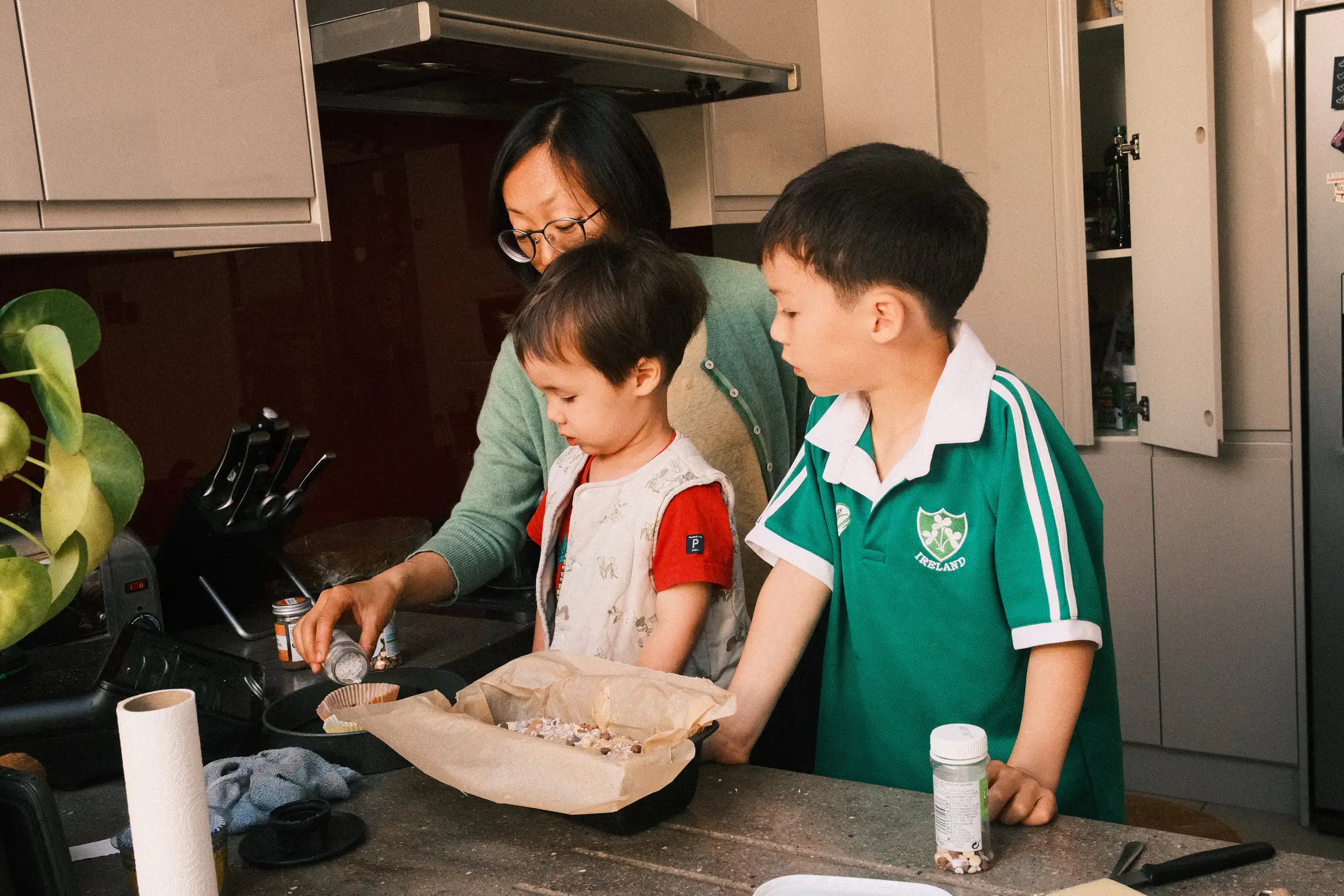 A mother and two boys in the kitchen. The two boys look on as the mother pours out sprinkles for the top of a cake.
