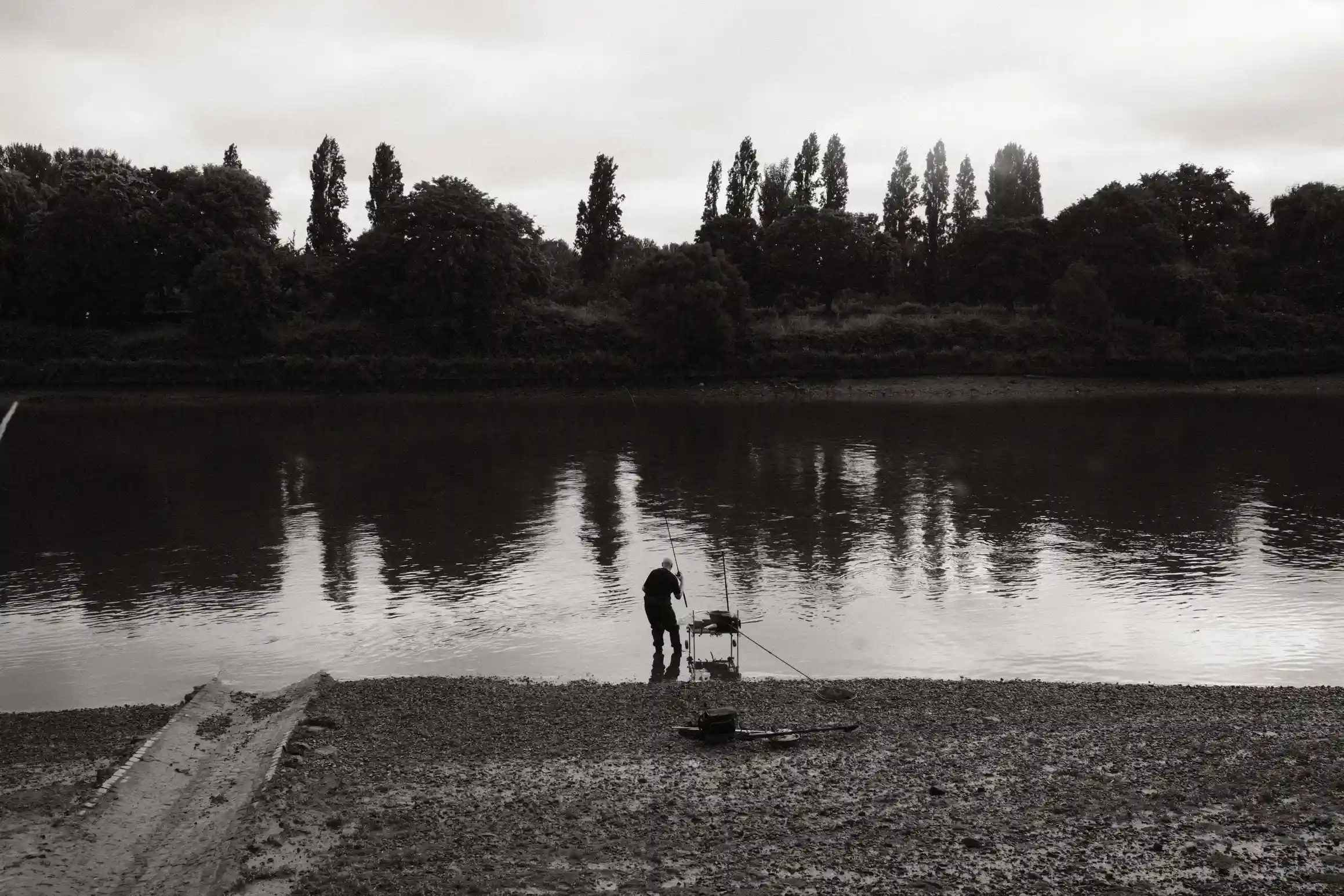 A black and white photo of a man of a man standing at the edge of a river fishing.