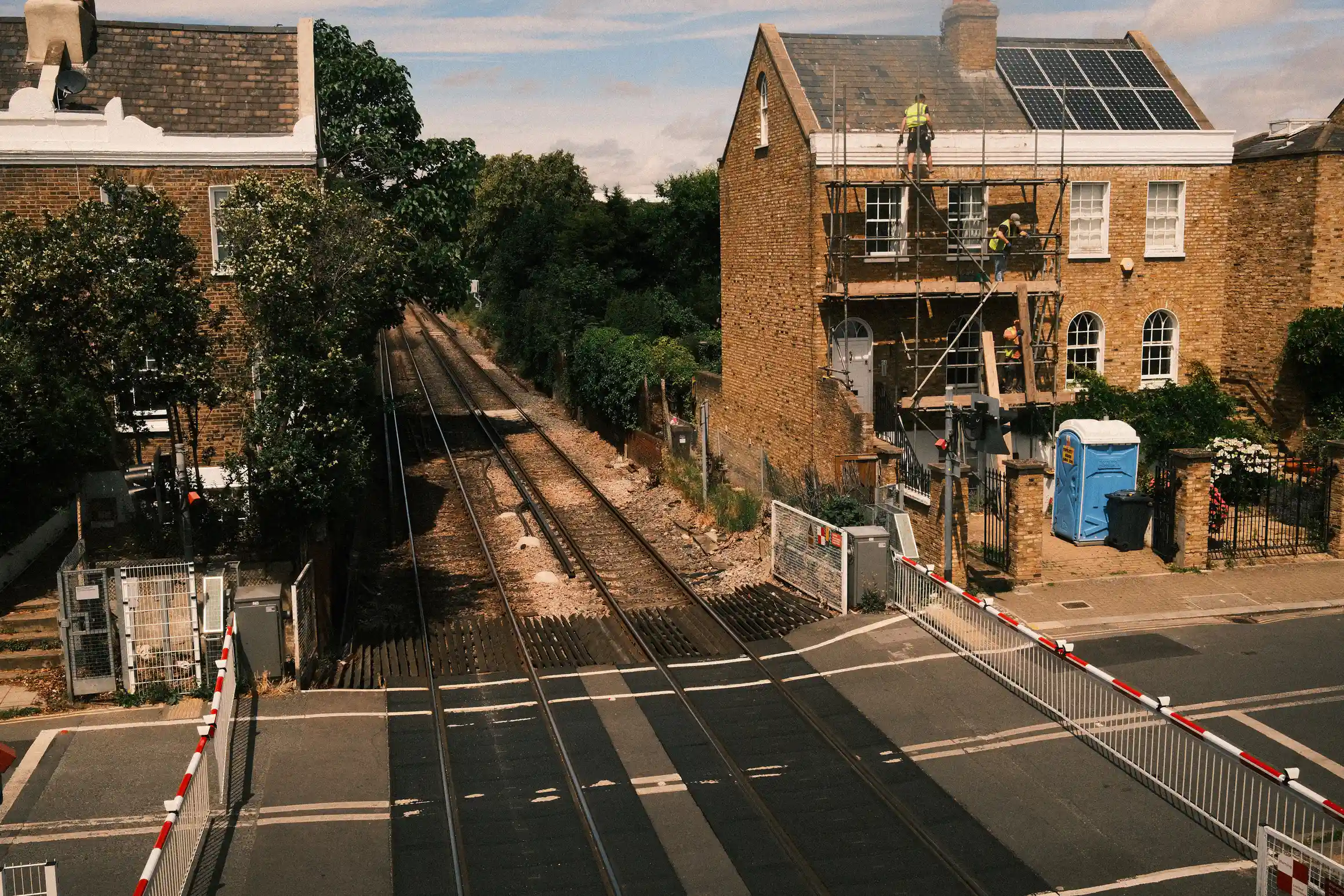 A photo looking down on a level crossing. The barriers are closed. On one side, workmen work on the roof of a house.
