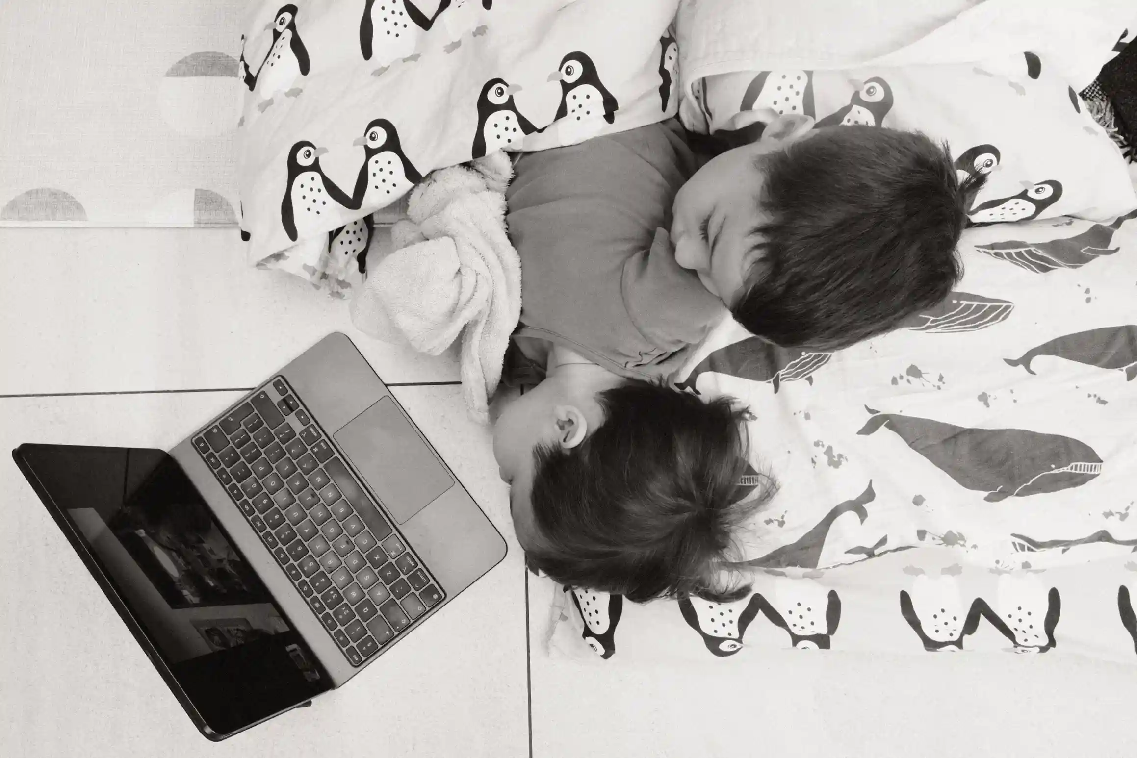 A black and white photo of two boys laying on the ground under a duvet looking at an iPad. 