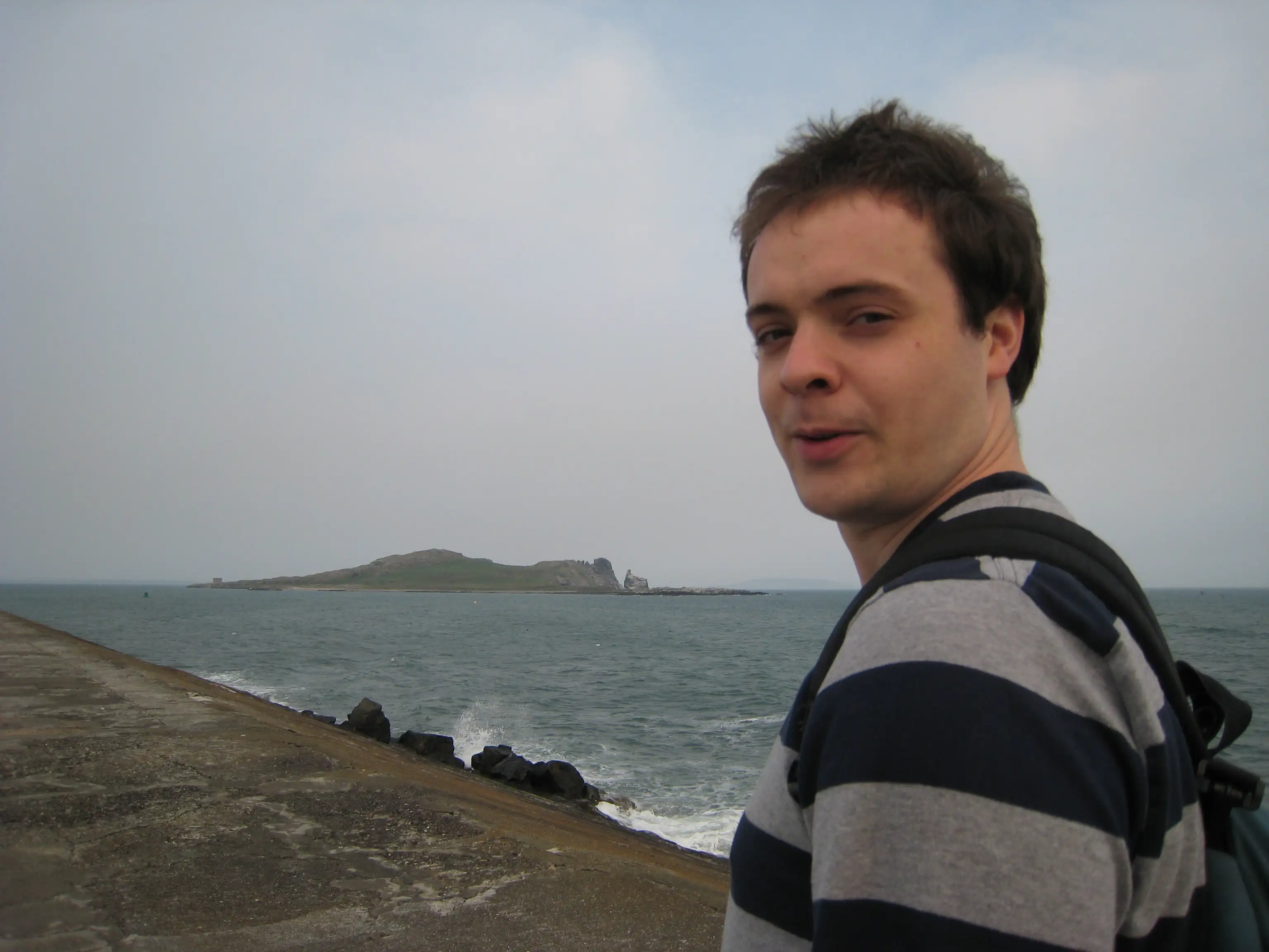 A photo of a person standing on a concrete pier, the ocean behind them. In the distance a small island. The person looks back towards the camera.