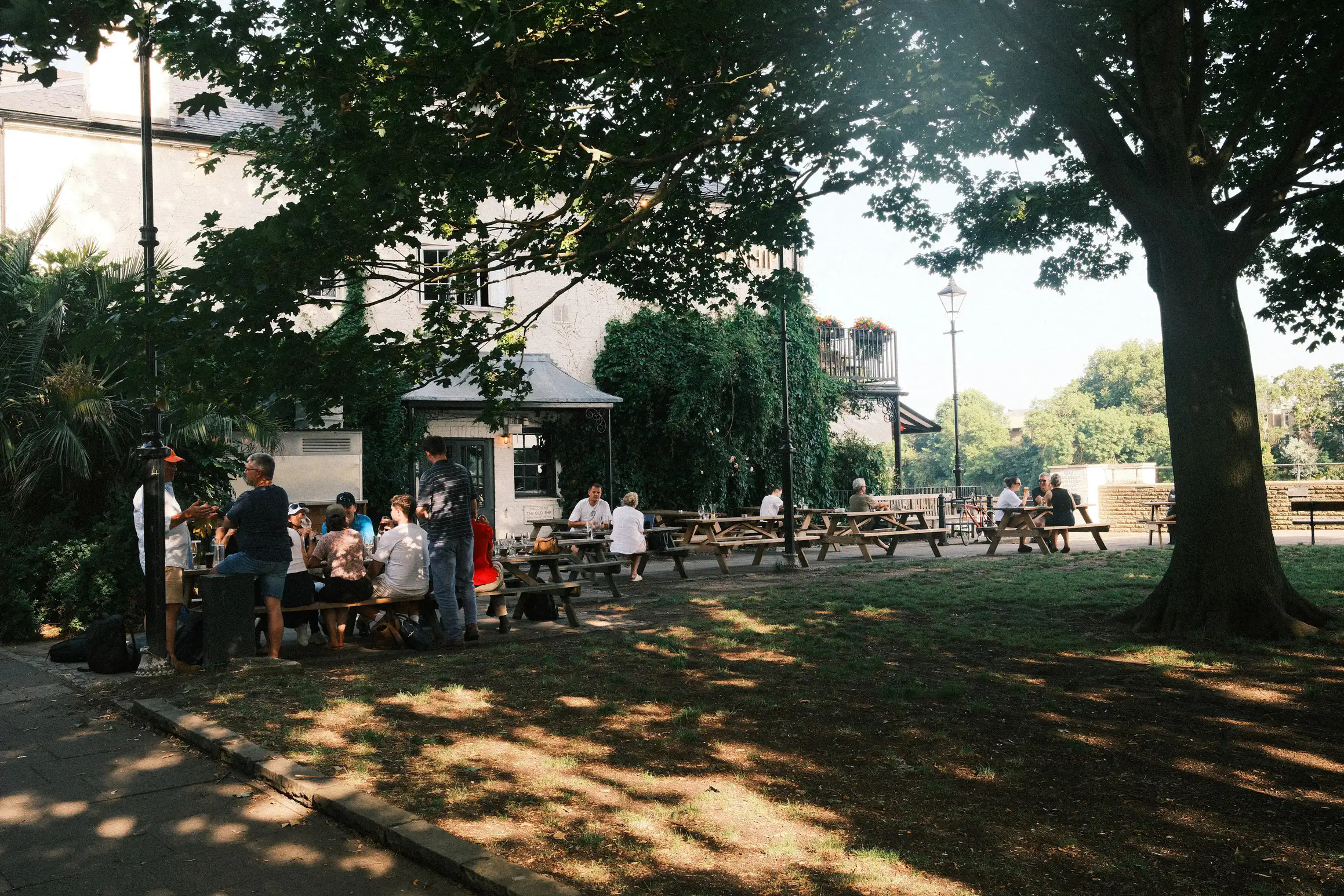 A shaded area outside a pub. In corner, group of people sit on a pub table. 
