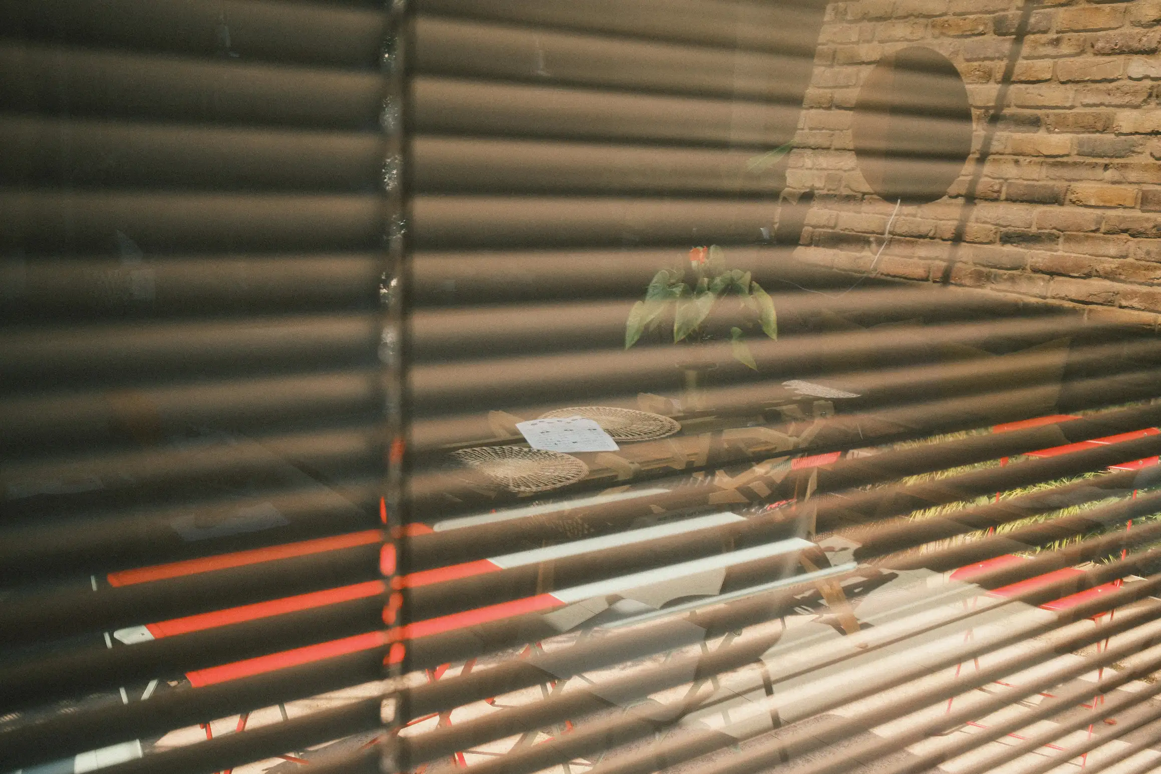 A photo of a partial reflection in a window. The gaps between the blinds allow light from outside to shine through, but the reflection of a table inside the room can also be seen in the glass.